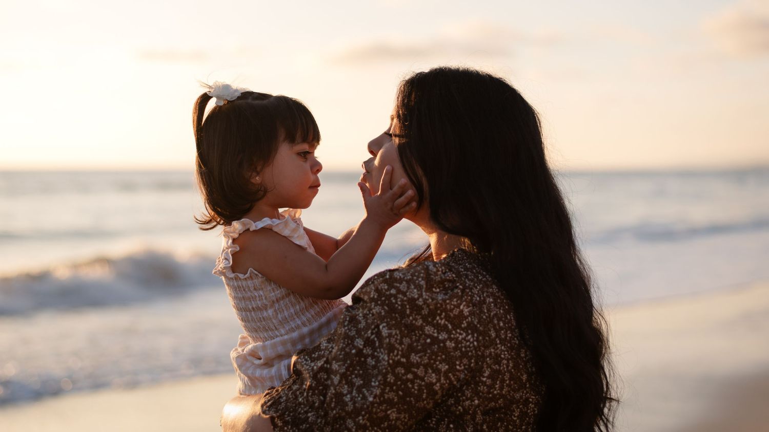 Mother holding toddler daughter on beach at sunset, sharing tender moment.