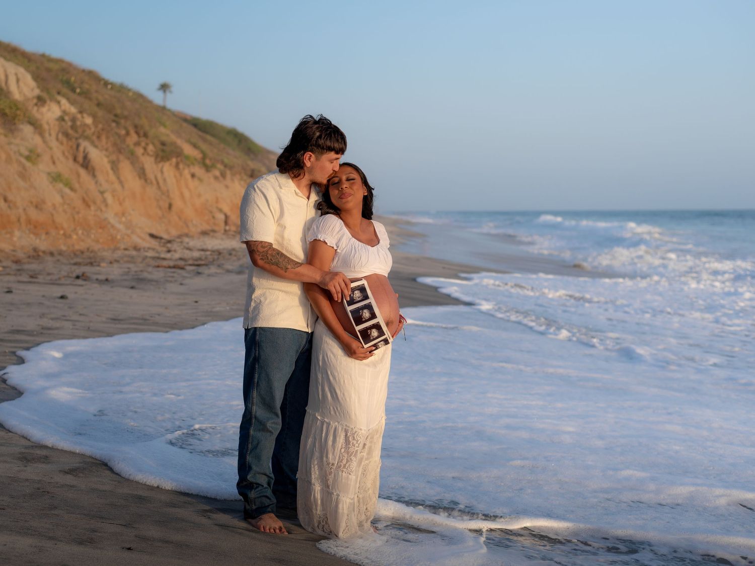 Couple embracing on a sandy beach at sunset, holding ultrasound photos during a maternity session.