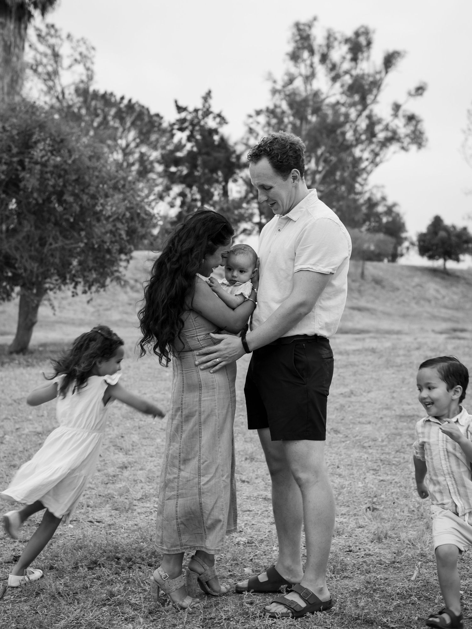 Black and white candid family photo of parents with newborn and two playful children outdoors.