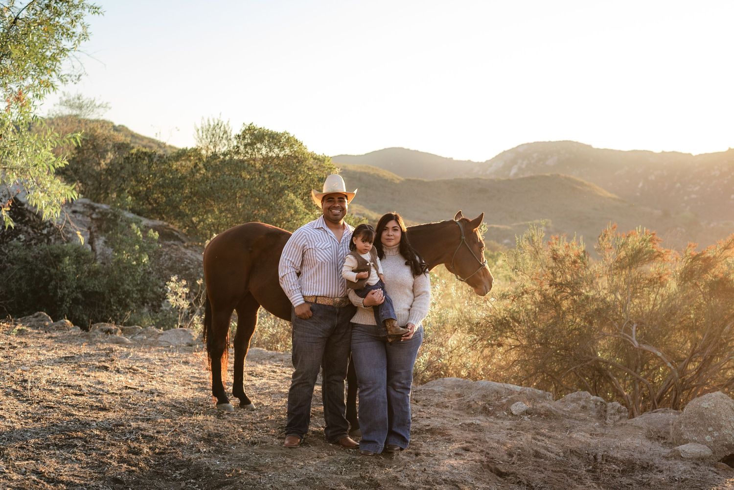 Family portrait with horse in sunlit mountain landscape at golden hour.