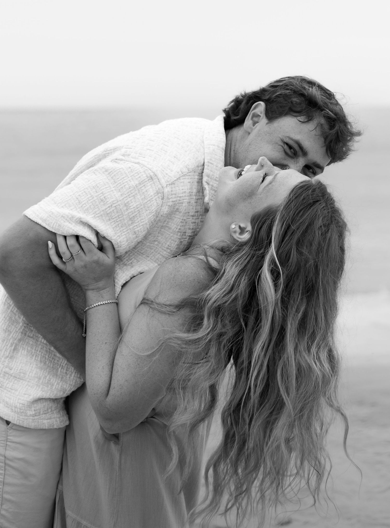 Black and white romantic photo of couple sharing a kiss on the beach, woman with long wavy hair.