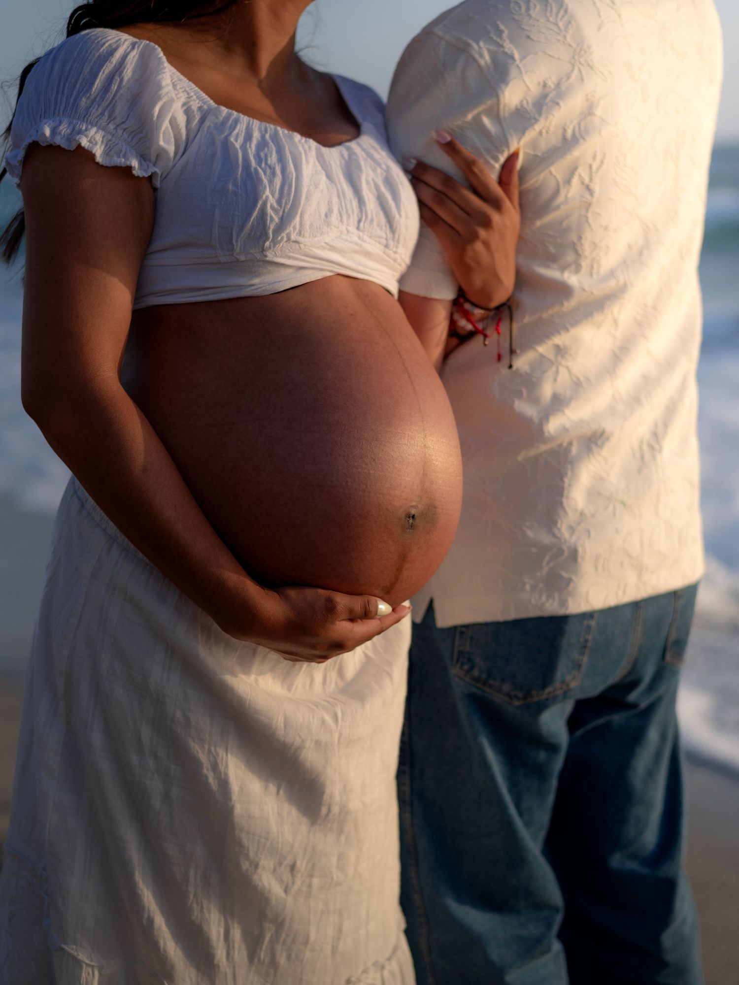 Close-up of a pregnant woman in a white dress cradling her bare baby bump on a beach at sunset.