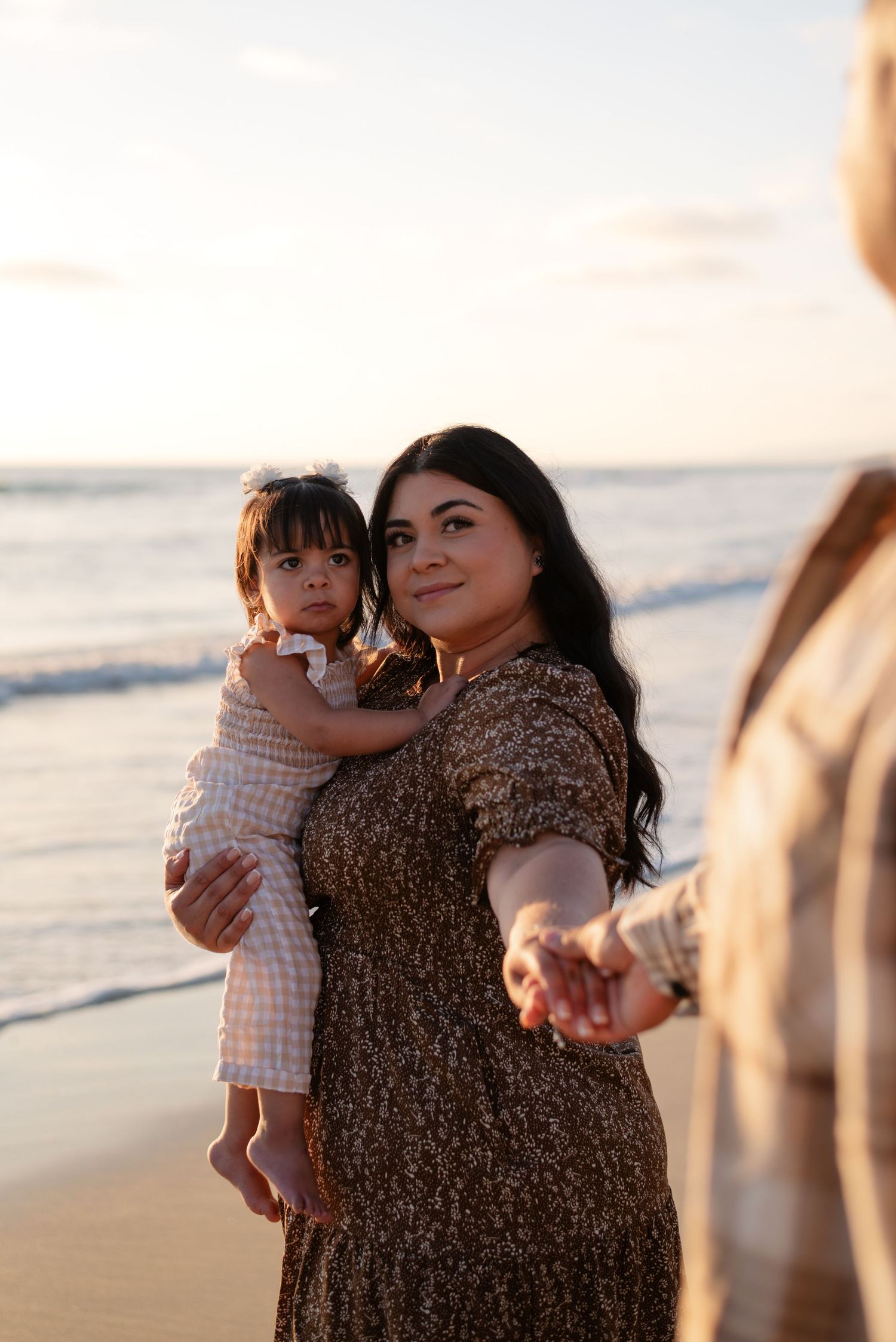 Mother holding her toddler daughter reaches out her hand toward someone on a sunset beach.