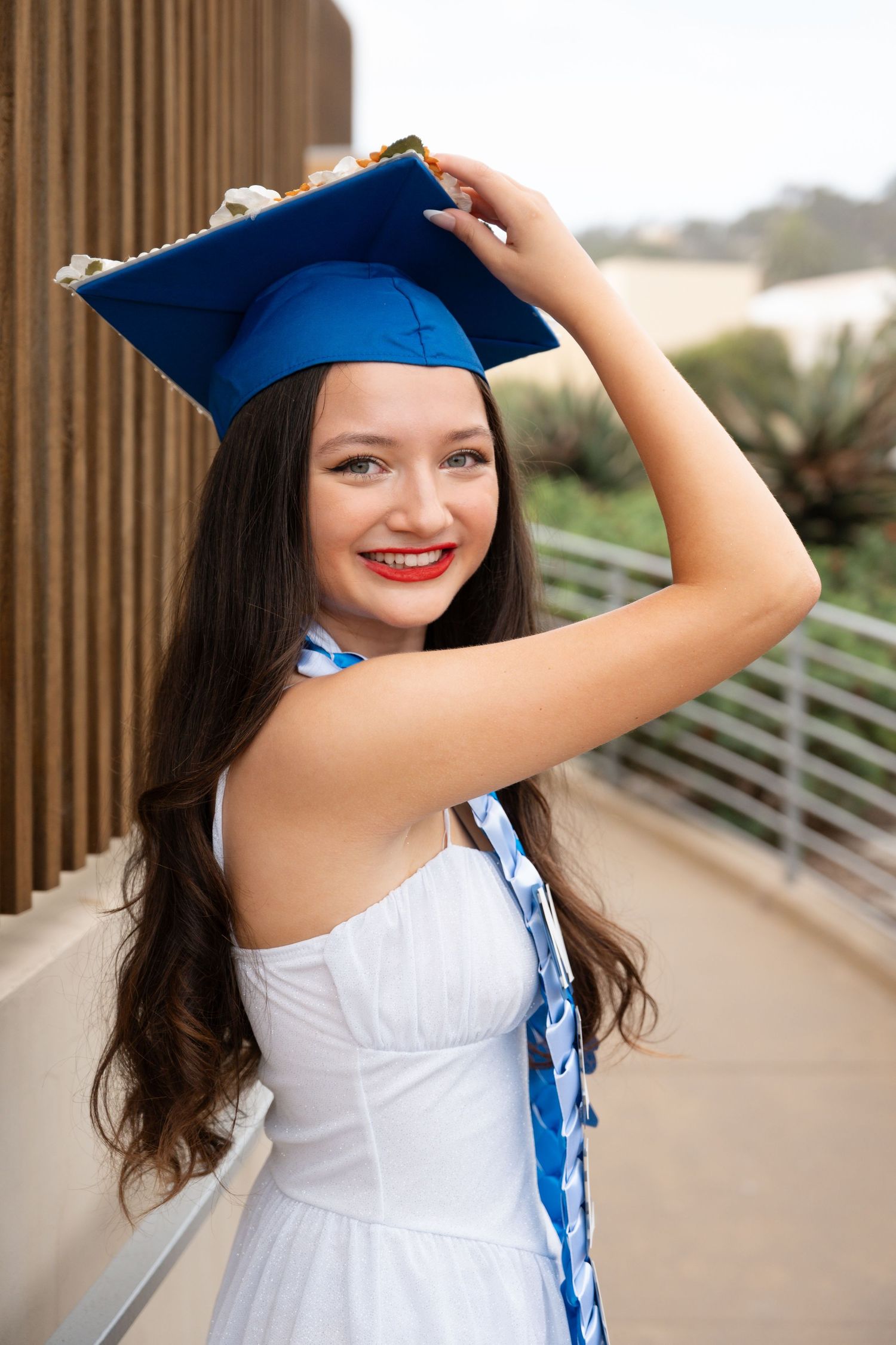Young woman in white dress and blue graduation cap smiles proudly, holding her mortarboard outdoors.