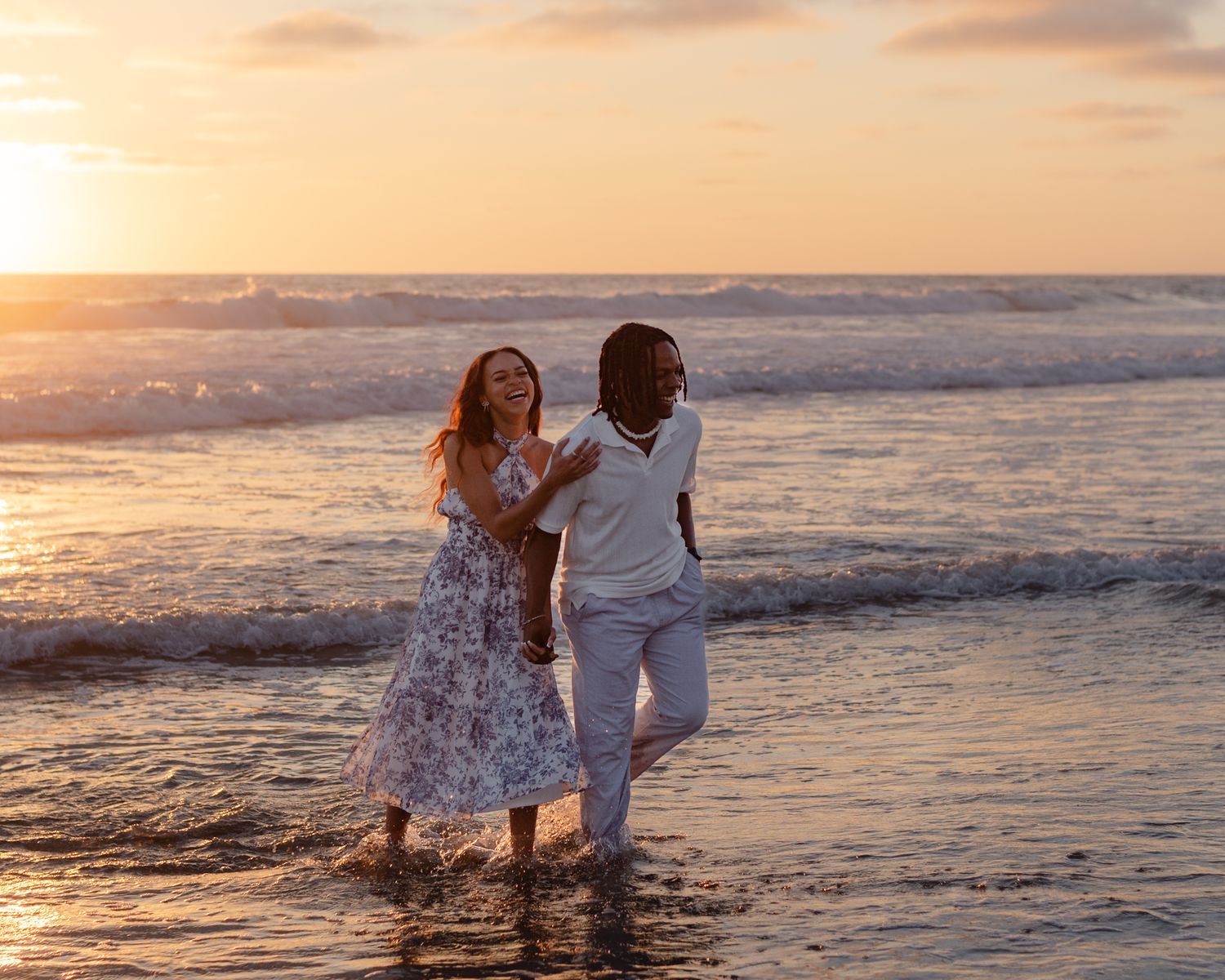 Couple laughing and splashing through ocean waves together during a vibrant beach sunset.