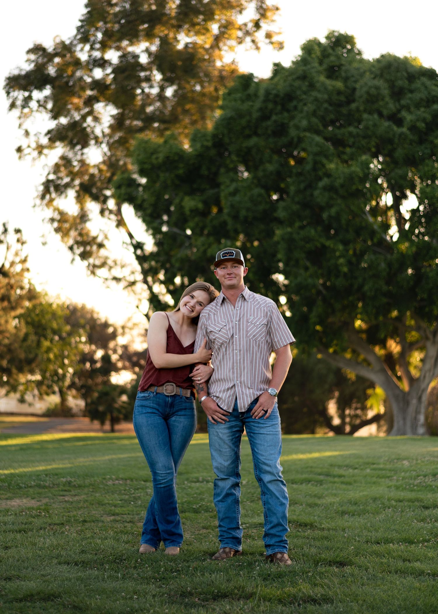 Couple posing in jeans on green lawn at sunset with large trees behind.