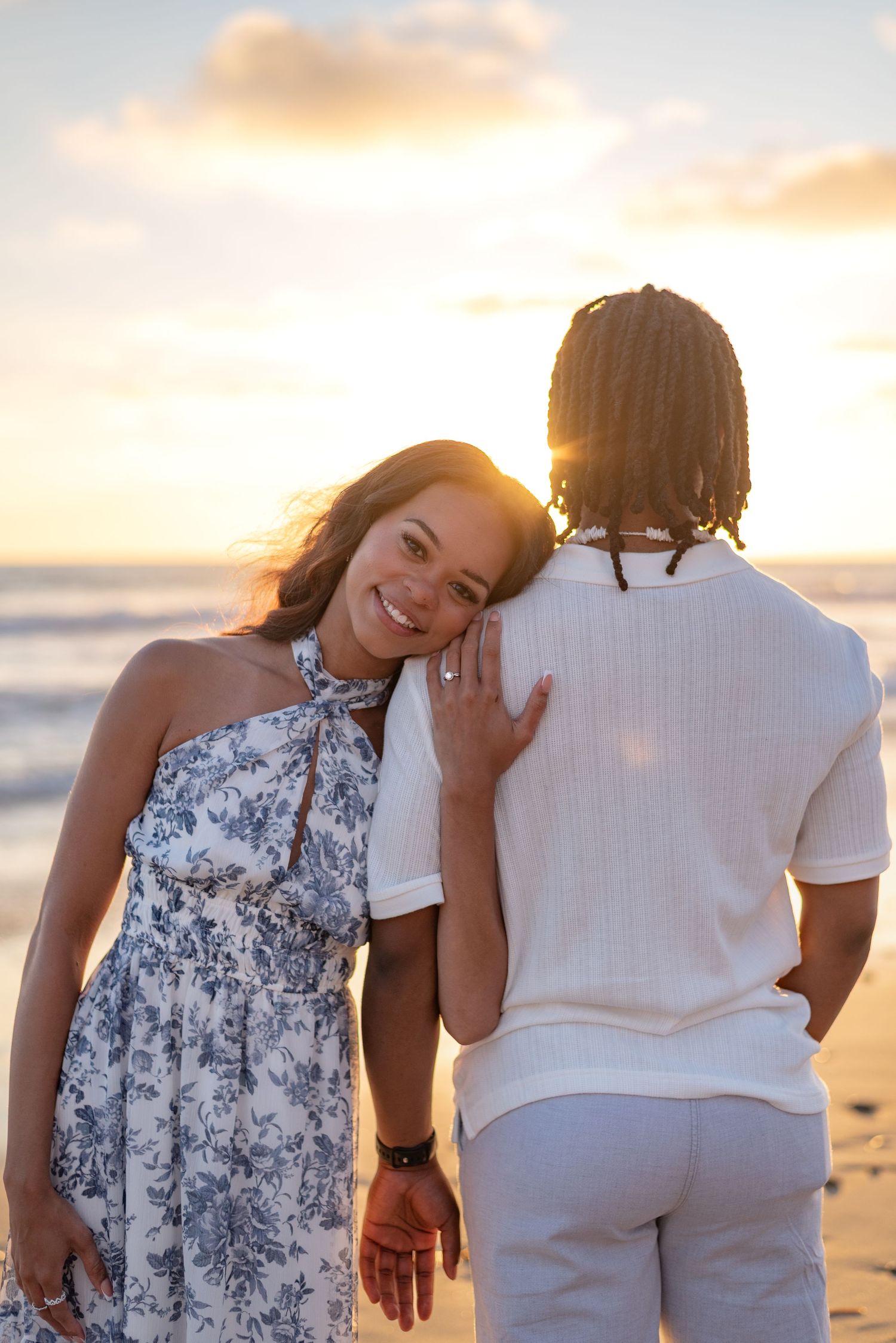 Smiling woman in floral dress leaning on partner's shoulder at beach sunset.