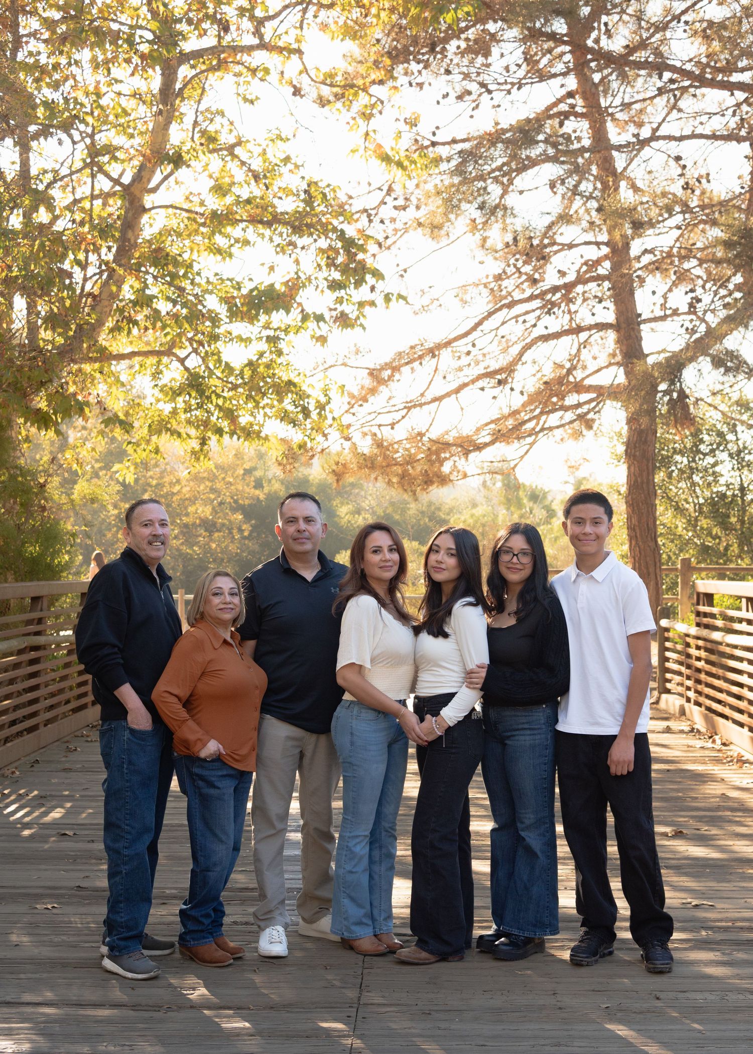 Extended family of seven posing on wooden bridge surrounded by autumn trees.