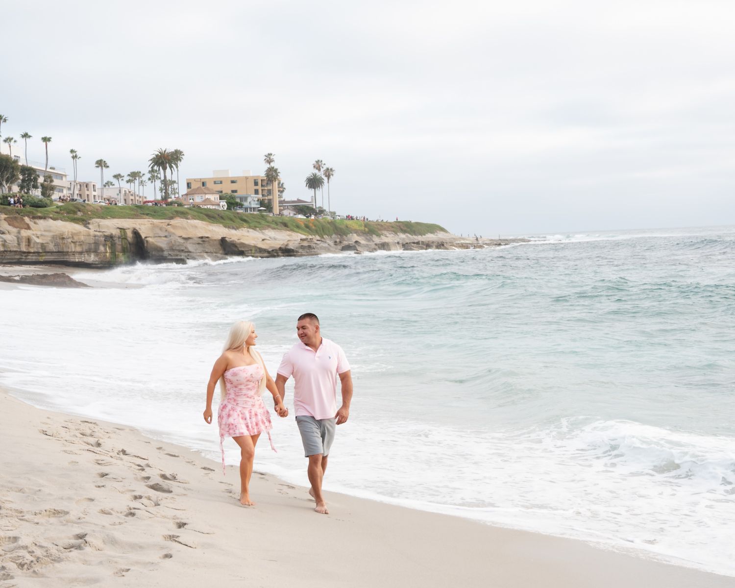 Couple holding hands walking barefoot on a sandy beach with ocean waves and palm tree-lined cliffs in the background.