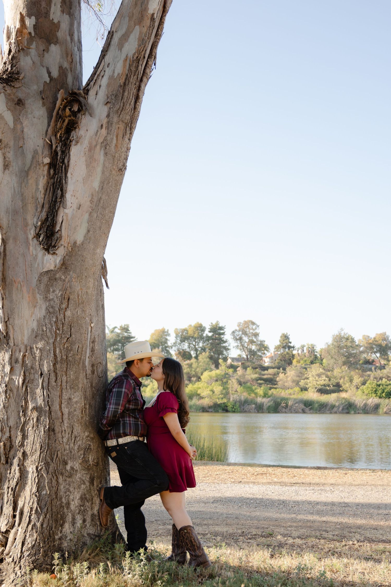 Couple sharing a romantic kiss leaning against a large tree by a river, woman in red dress and man in cowboy hat.