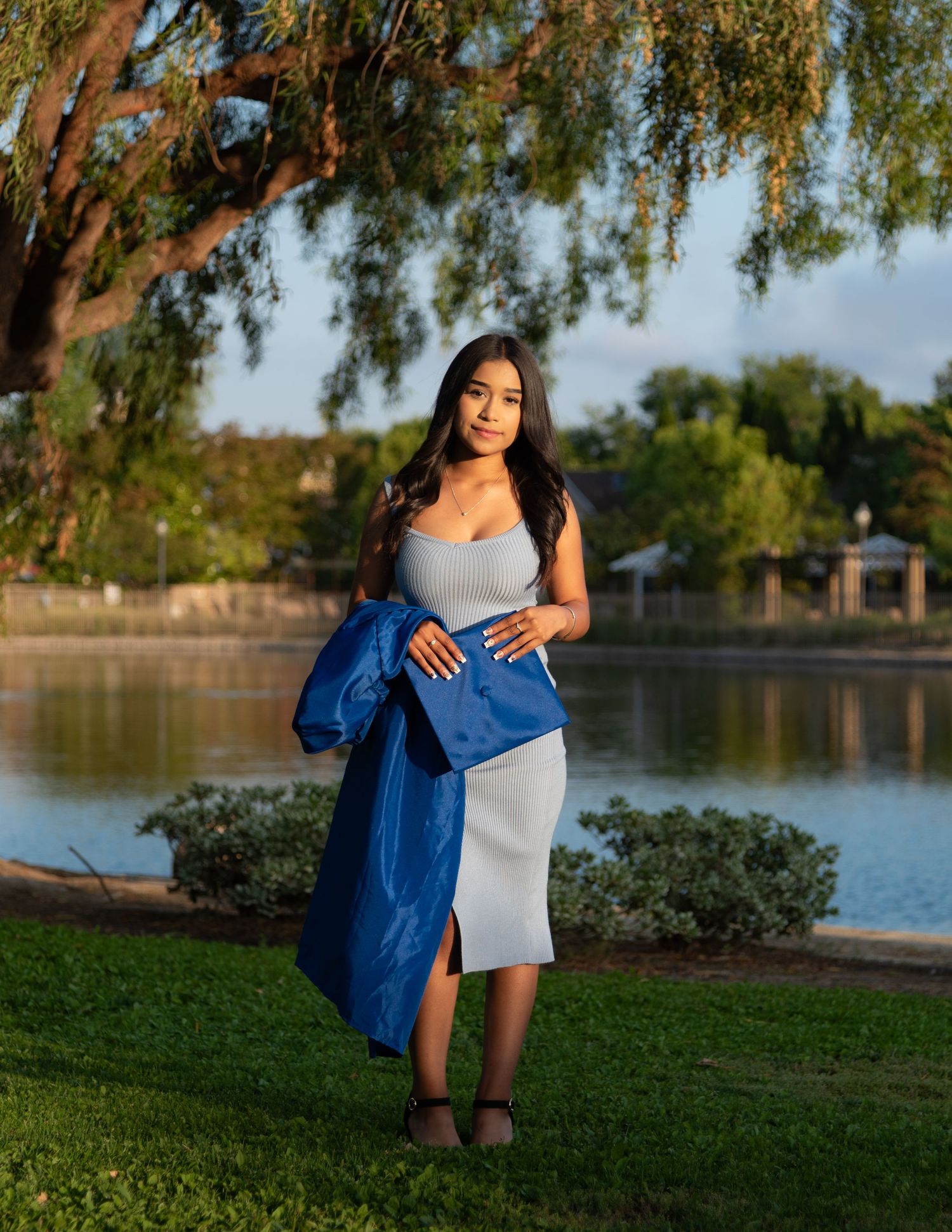 Graduate in blue robe and gray dress stands by sunlit lake for senior portrait.