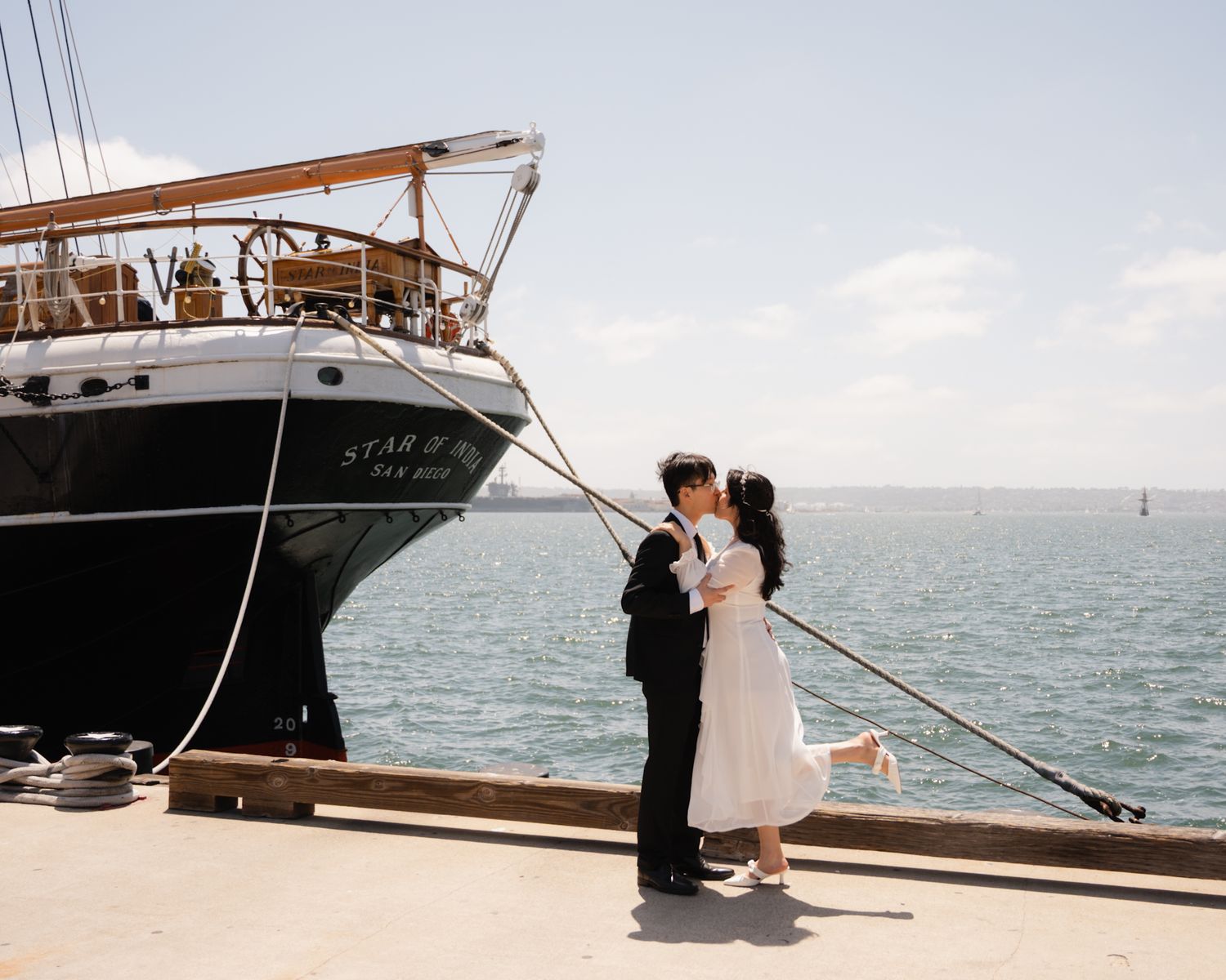Couple sharing a kiss on a dock beside a tall ship on a sunny day.