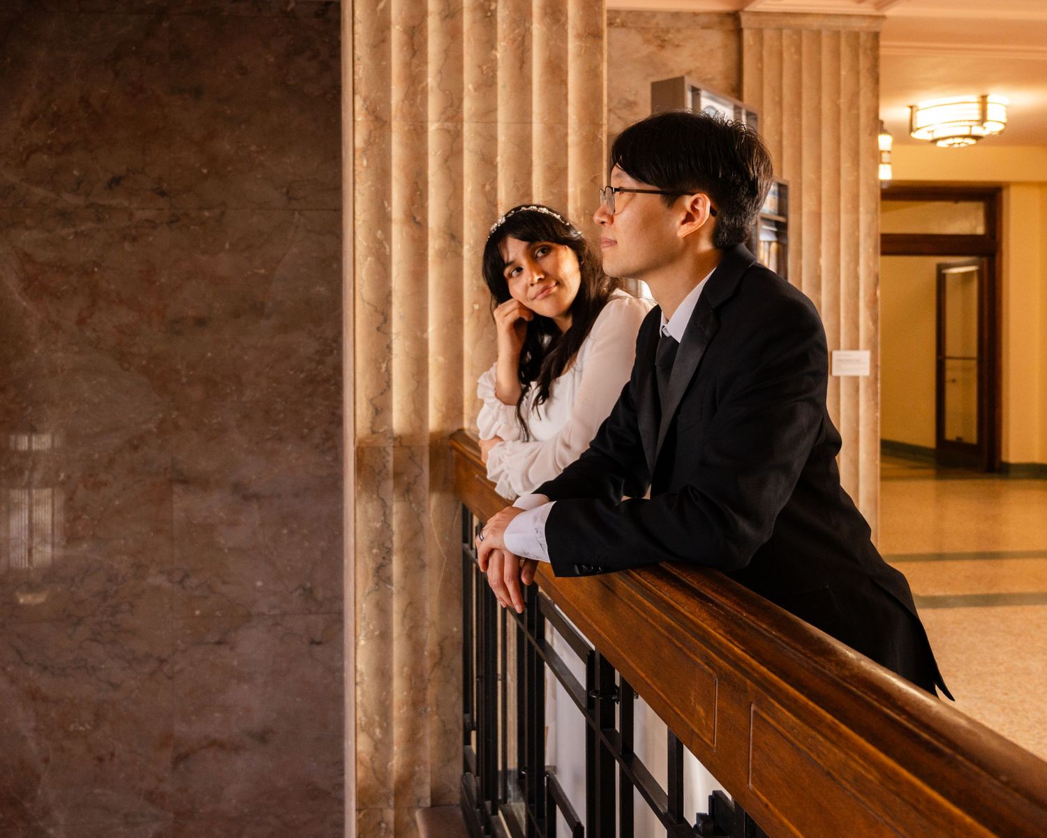 A couple in formal attire leans on a railing in an elegant marble hallway with ornate columns.