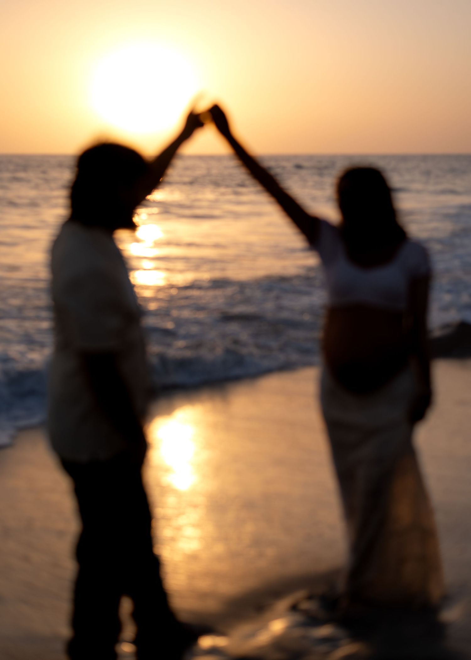 Couple holding hands forming heart shape silhouetted against glowing orange beach sunset.