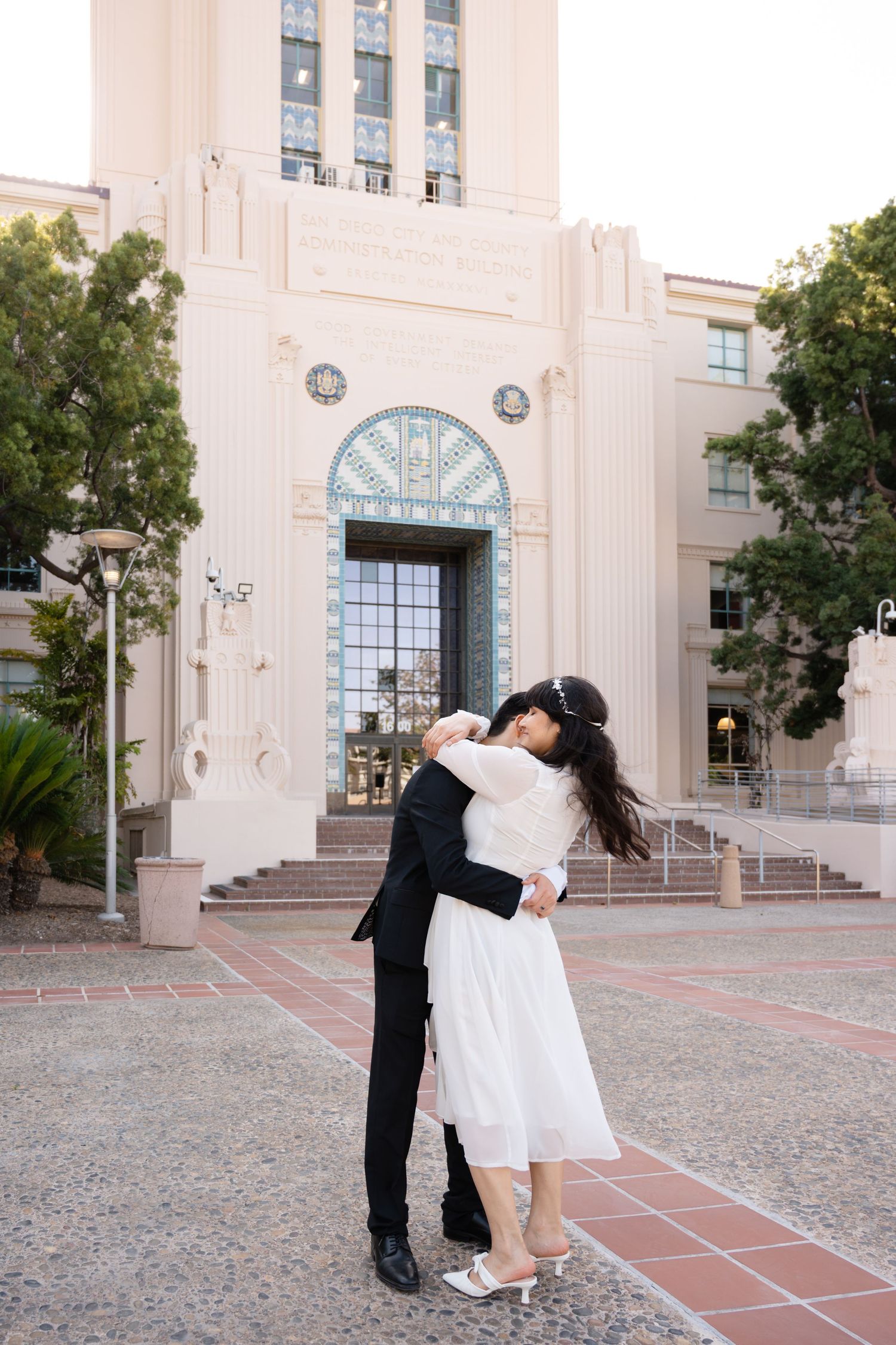 Newlywed couple sharing a romantic embrace outside an Art Deco building in a sunny courtyard.
