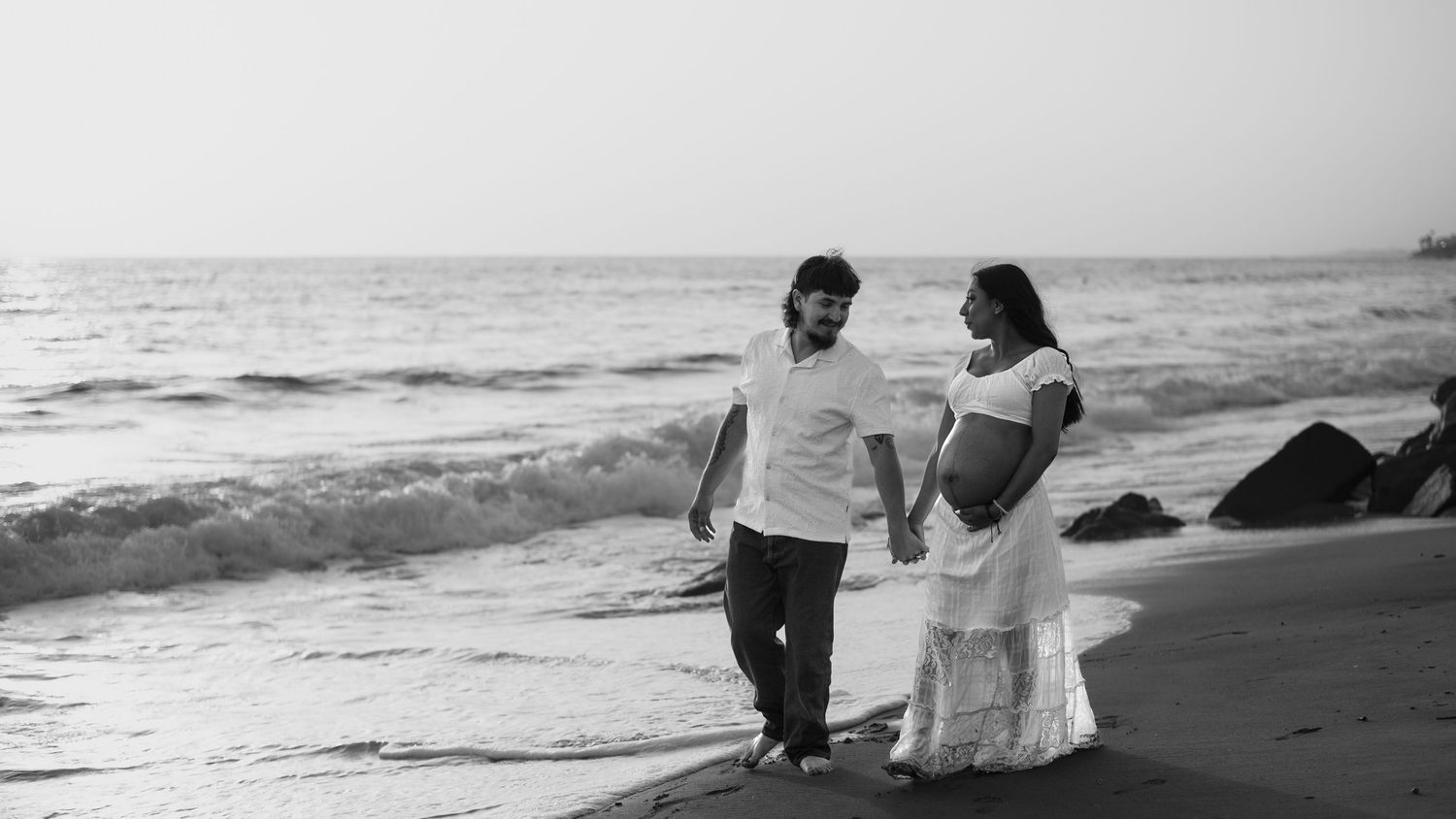 Couple walks hand in hand along ocean shore in black and white maternity photo.