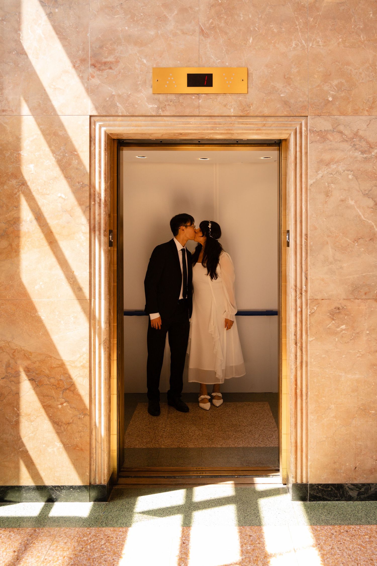 Couple sharing a kiss in an elegant elevator doorway, bride in white dress and groom in dark suit.