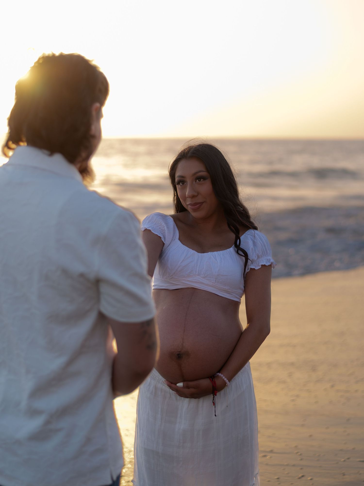 Pregnant woman in white dress holding baby bump at sunset beach, facing her partner in a maternity photo session.