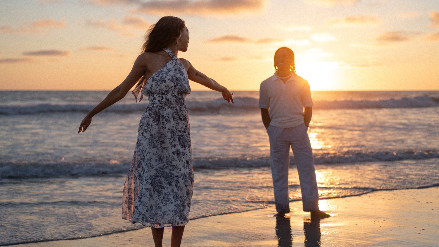Two women enjoying a romantic sunset on a tropical beach, one in a floral dress with arms outstretched.