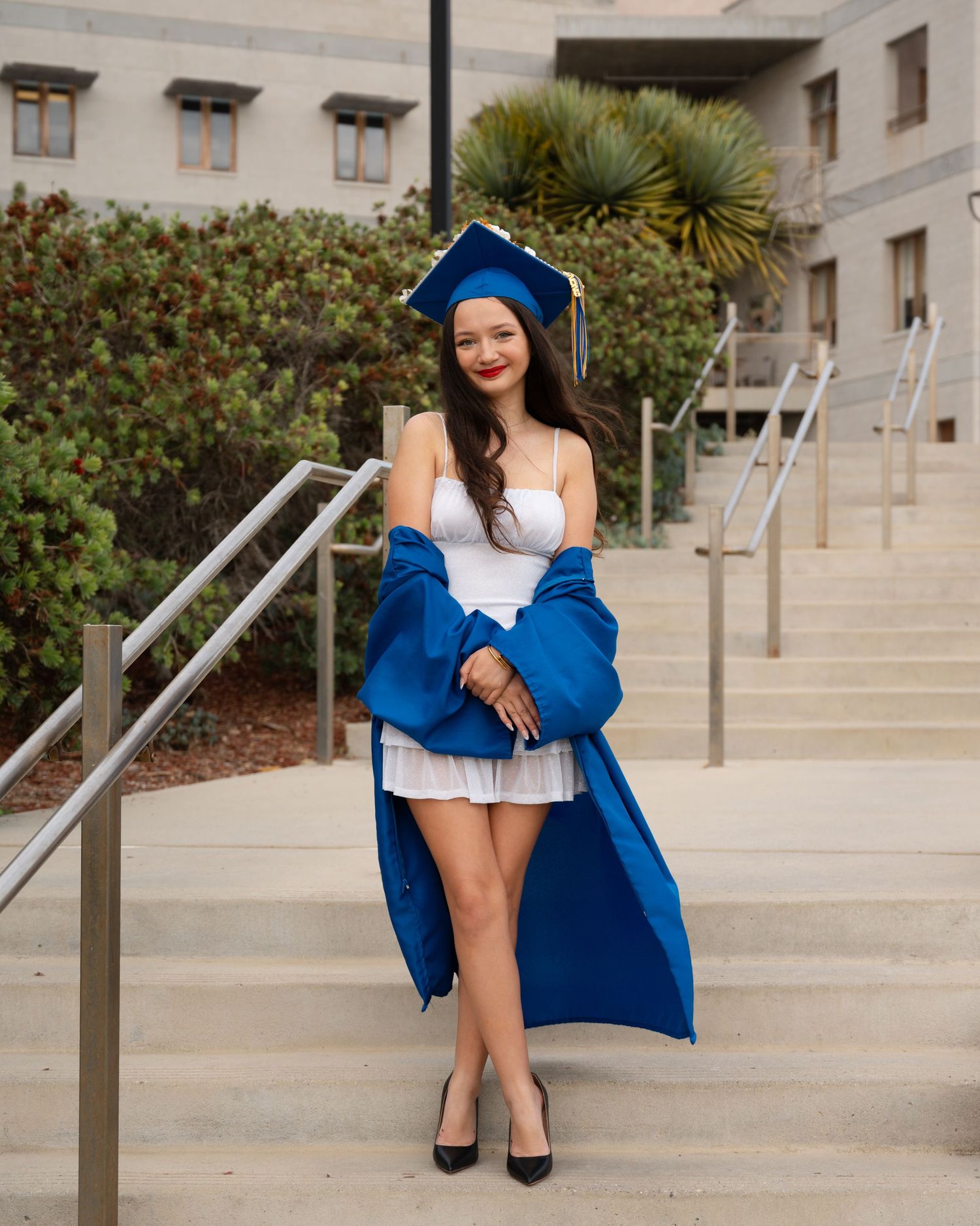 Young woman in blue graduation cap and gown over white dress, posing on steps at university campus.