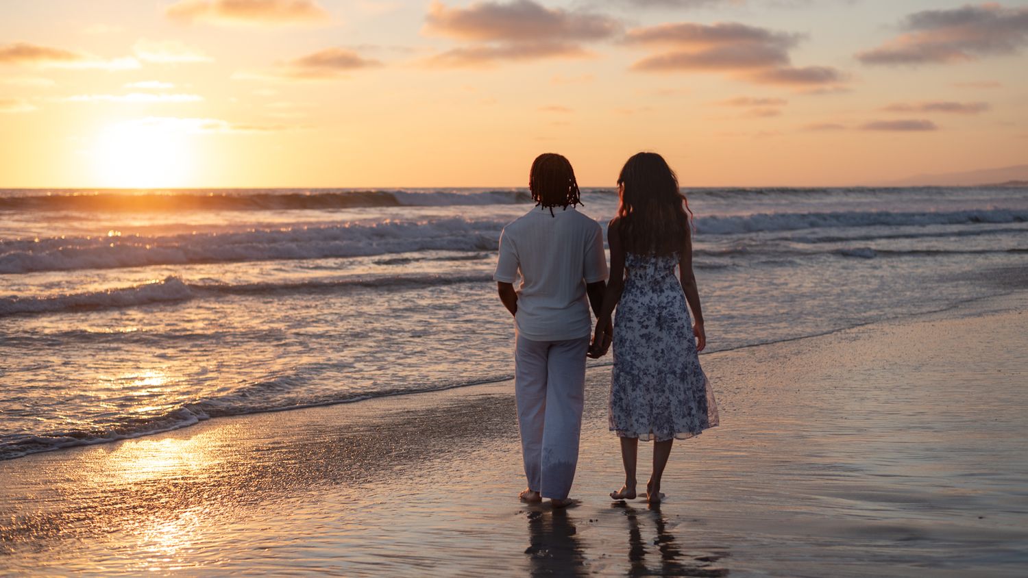 Two women holding hands walking barefoot along a reflective beach shoreline at a beautiful golden sunset.