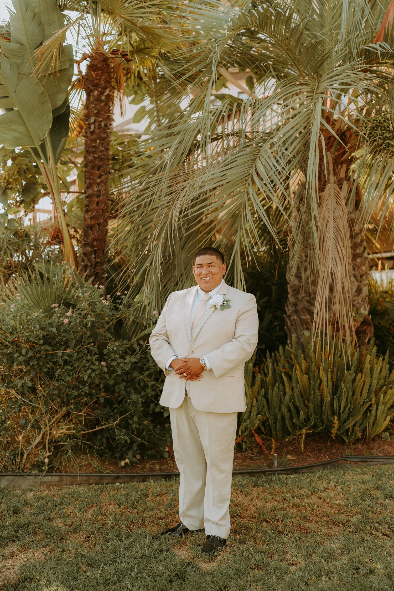 Groom in cream suit stands confidently before lush tropical palm trees at outdoor wedding venue.