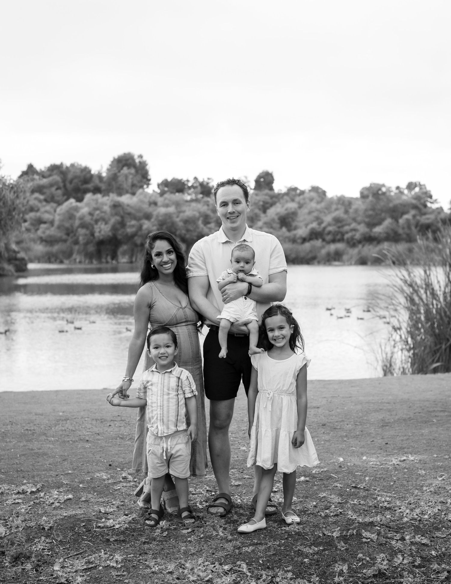 Black and white family portrait of parents with three young children near a peaceful lake.
