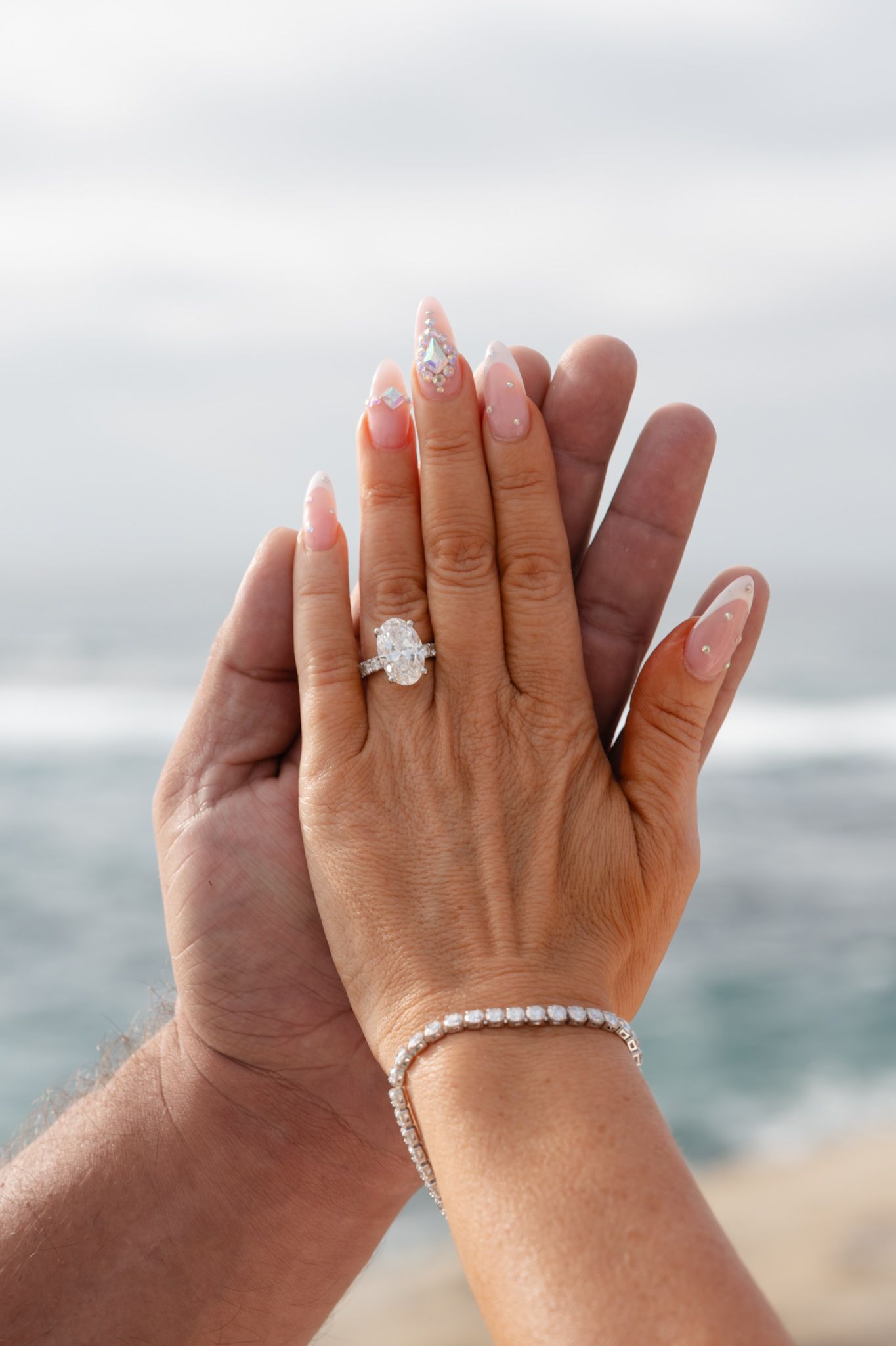 Two hands pressed together showing a large oval diamond engagement ring and tennis bracelet against an ocean backdrop.