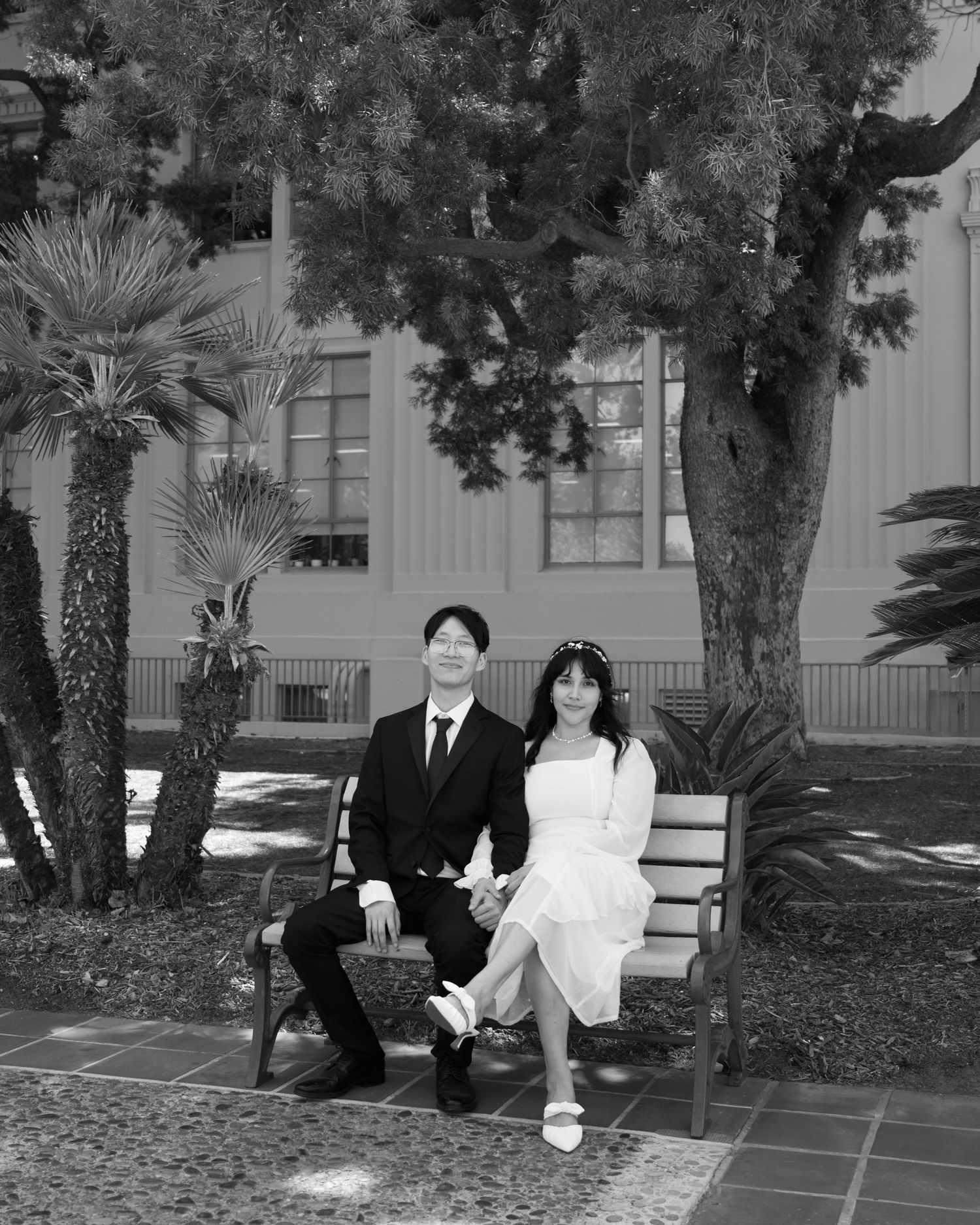 Couple in wedding attire sitting on a bench outdoors, black and white photo with trees and building.