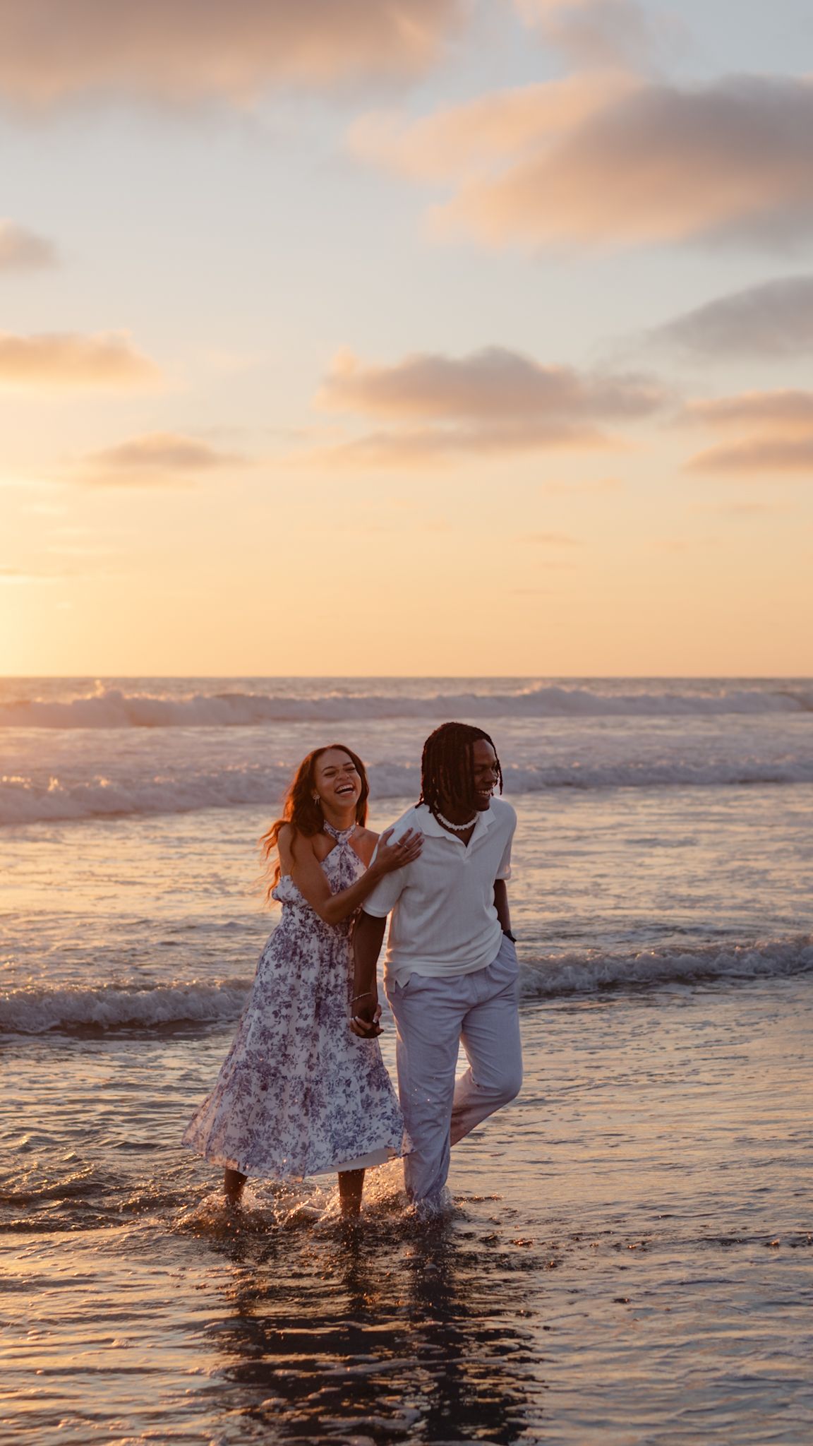 Laughing couple splashing in ocean waves at sunset, woman in floral dress and man in white outfit on beach.