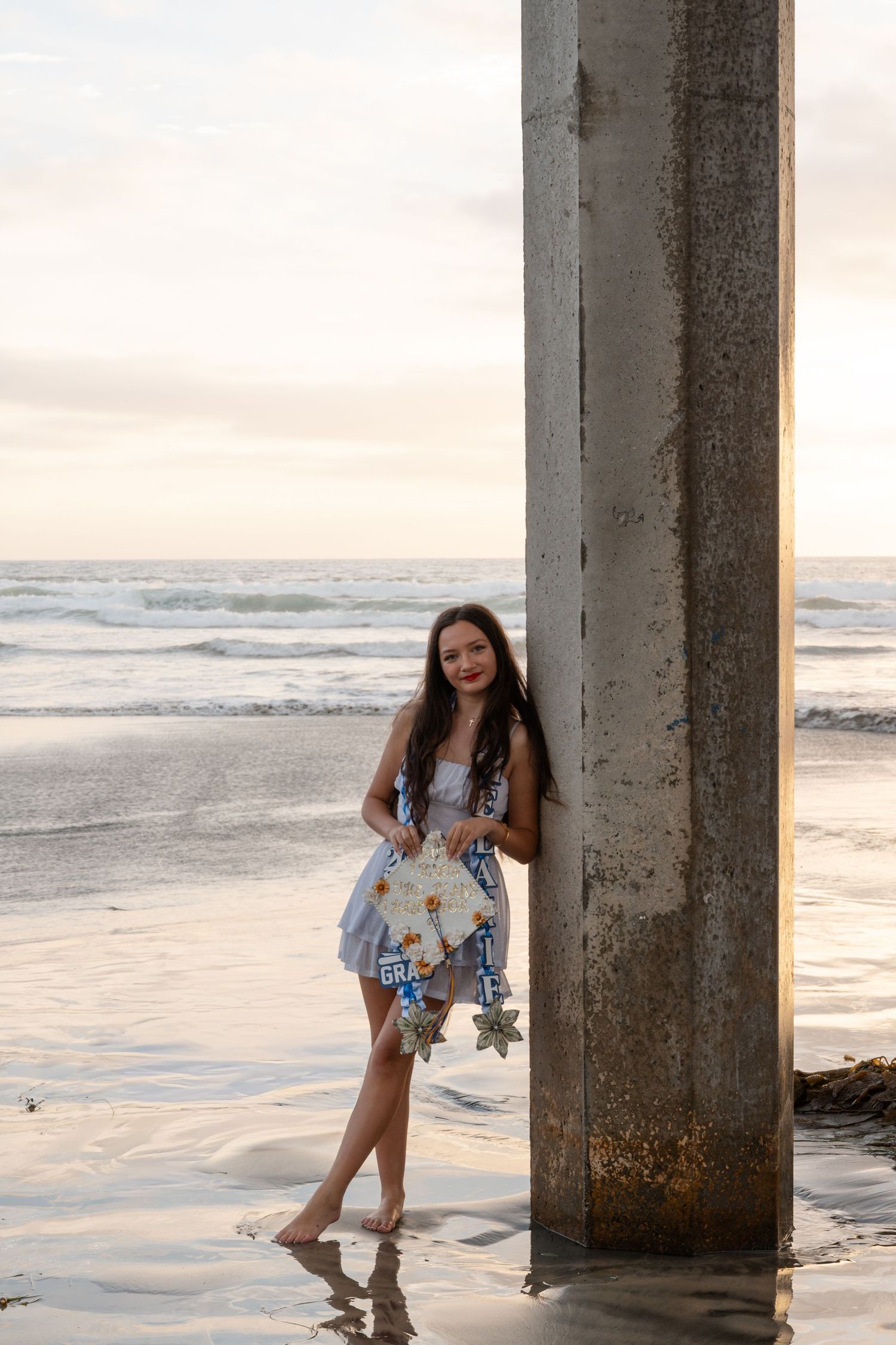 Young woman in floral dress leans against a concrete pier on a sandy beach at sunset with ocean waves behind her.
