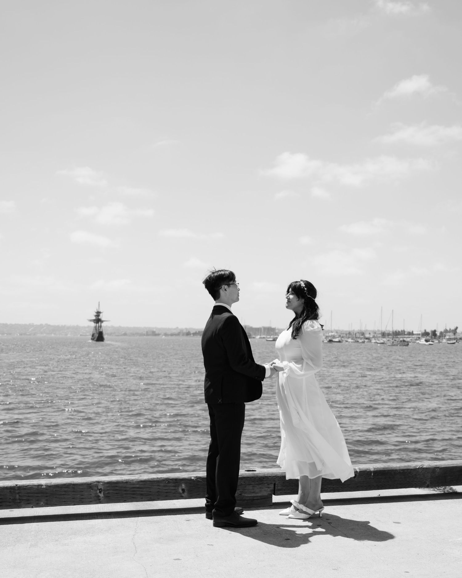 Black and white photo of a couple in wedding attire holding hands by the waterfront on a sunny day.