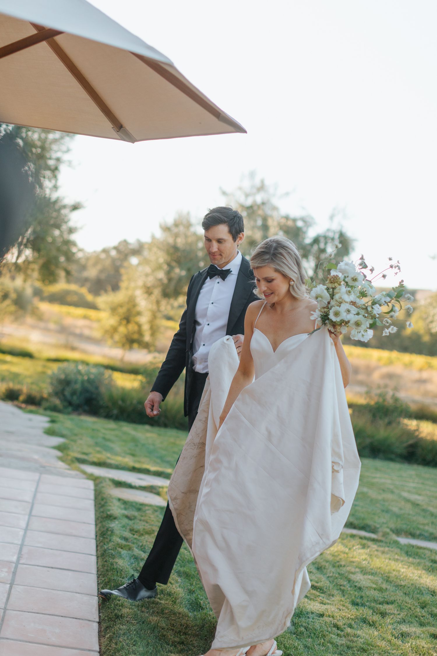 A bride and groom walk together under a patio umbrella in an outdoor garden setting.
