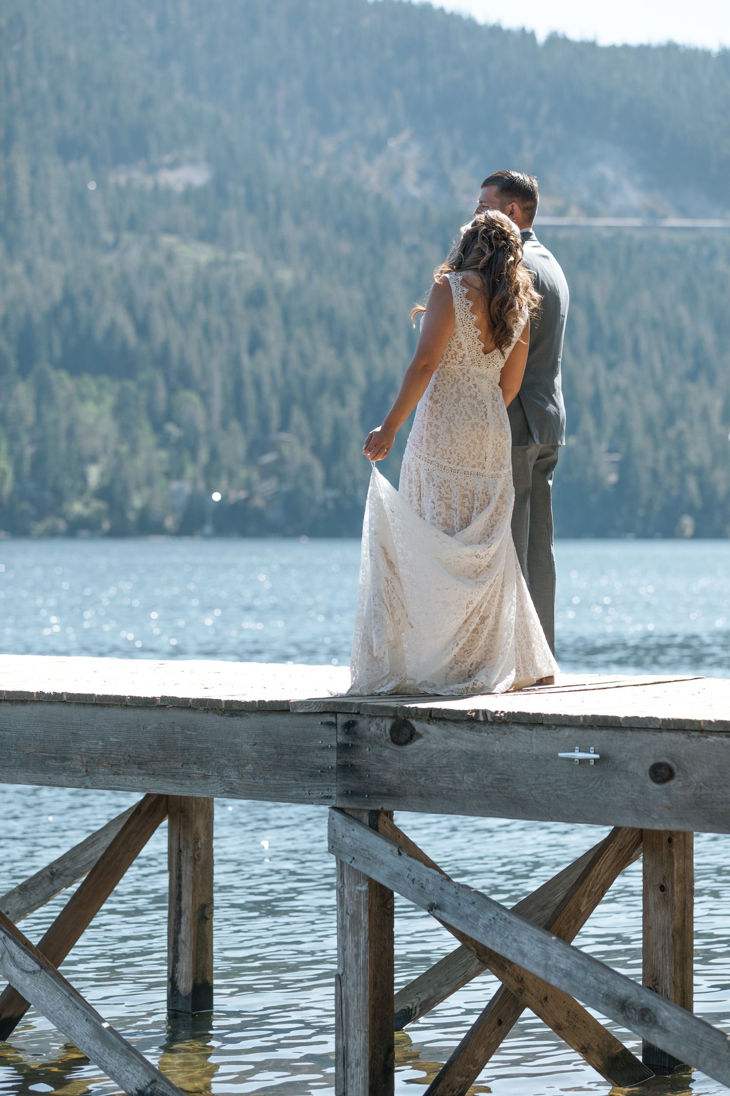 A couple sits together on a wooden dock overlooking a serene lake surrounded by evergreen-covered mountains.