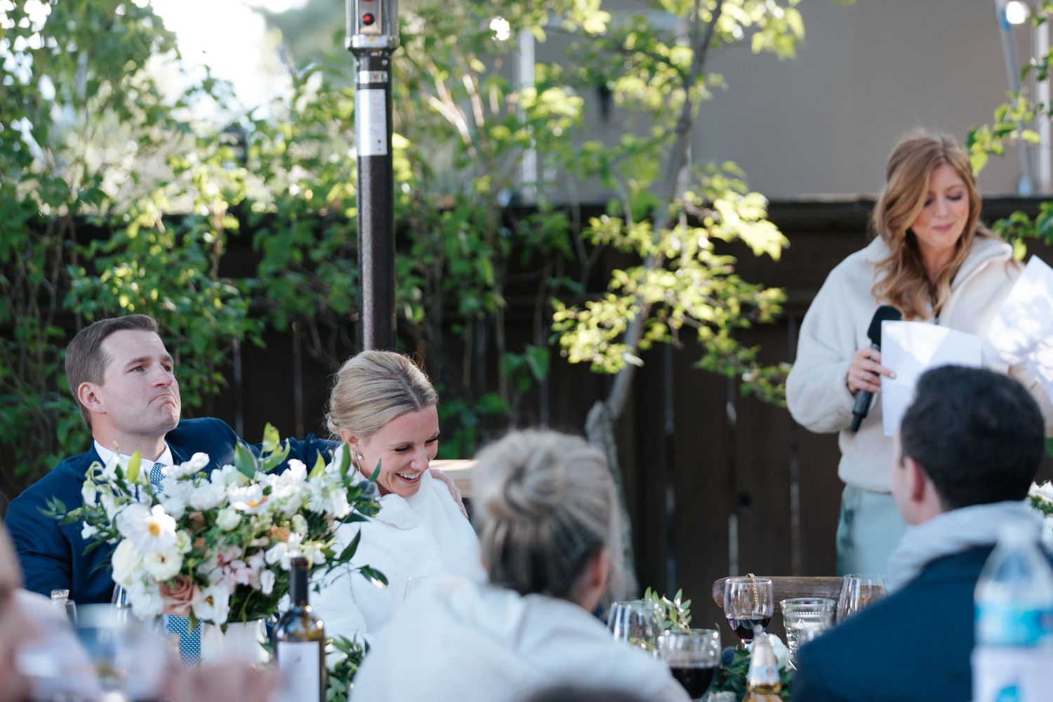 Wedding guests laugh and socialize during an outdoor reception under string lights and trees.