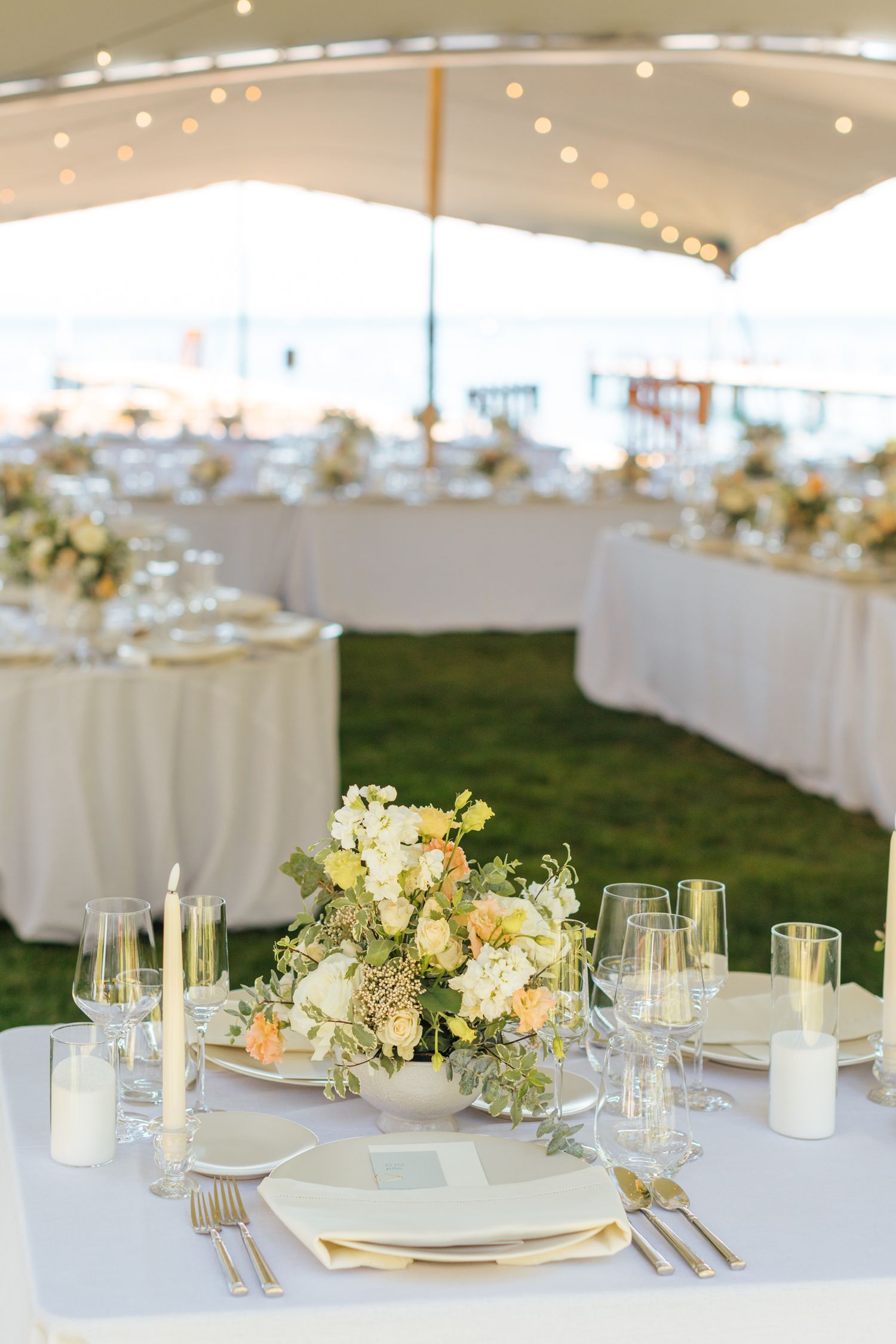 Elegant outdoor wedding reception under a white tent with floral centerpieces, candles, and gold flatware on white tables.