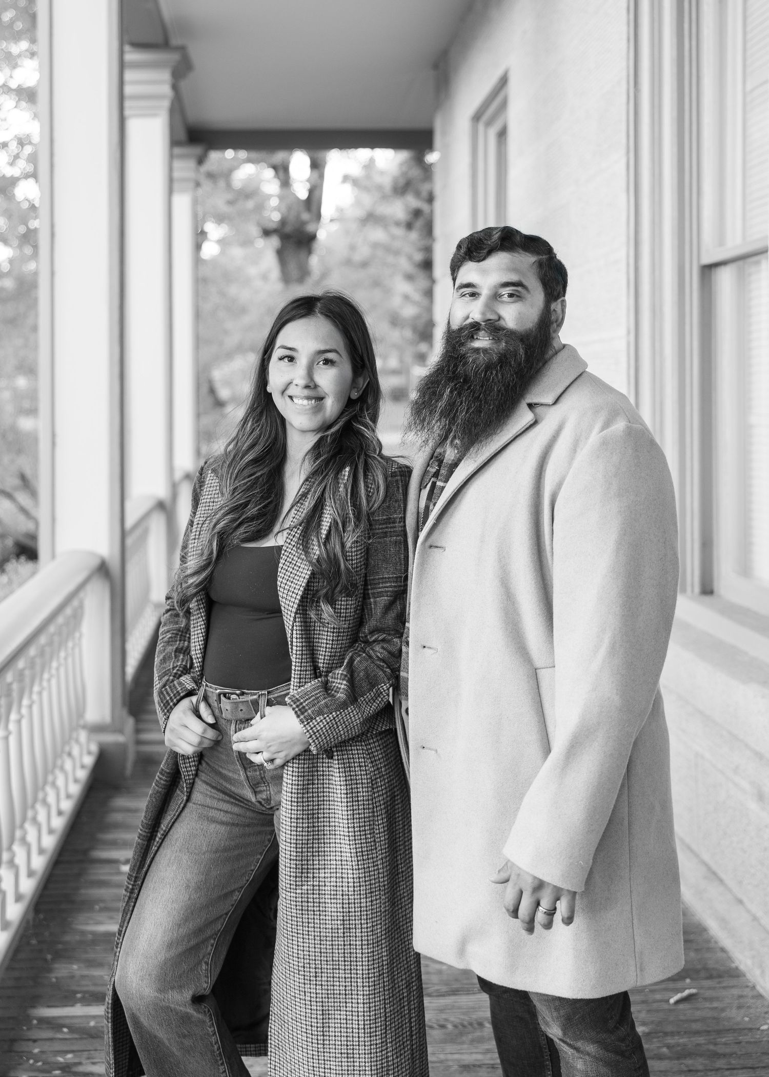 Professional photography duo posing on a white porch in matching beige coats.