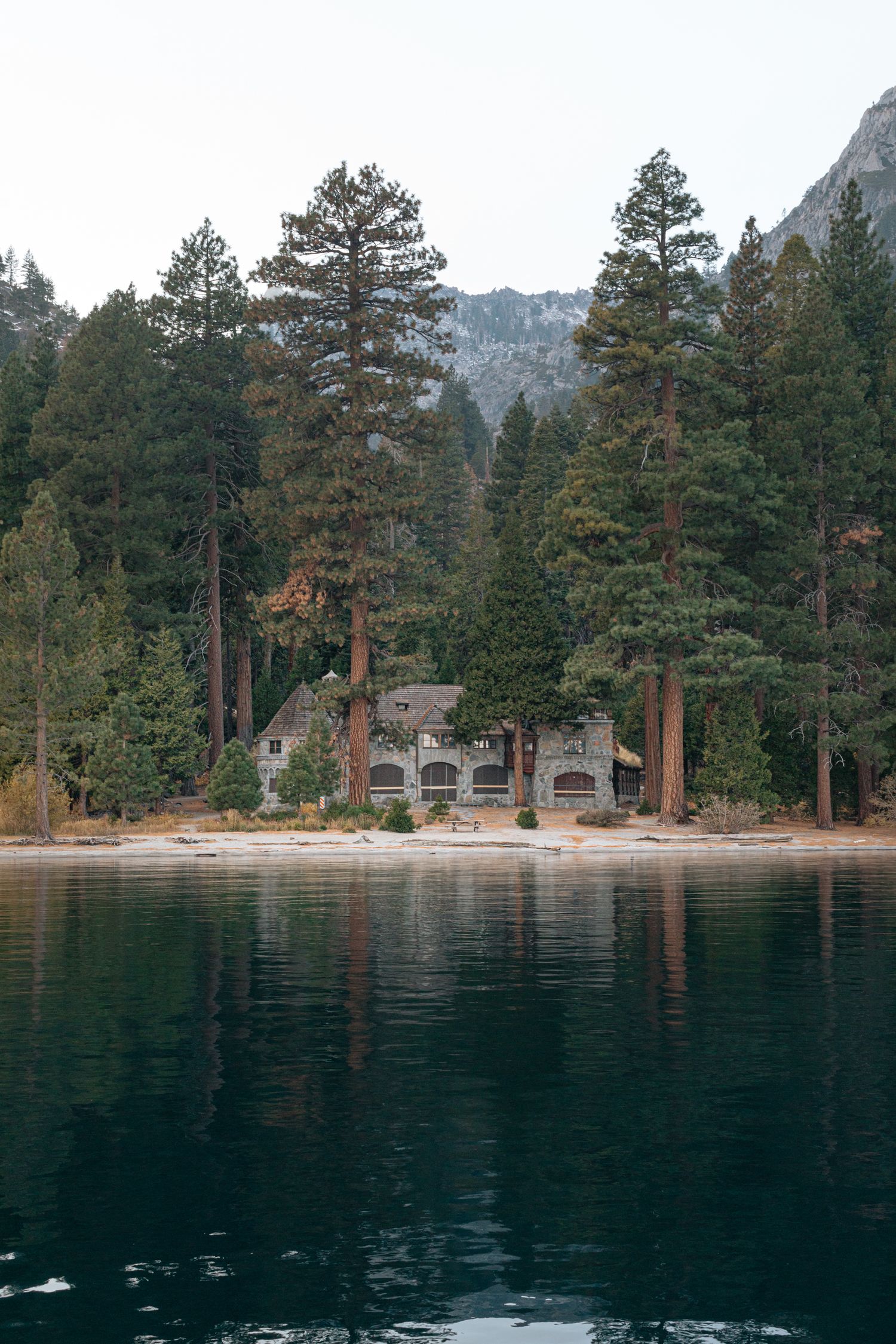 Lakeside view of pine trees and stone buildings along a mountain shoreline.