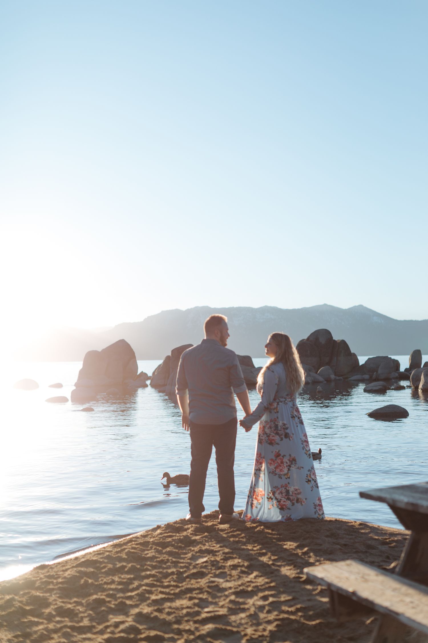 Silhouetted couple sits on beach with mountain and ocean views at golden hour.