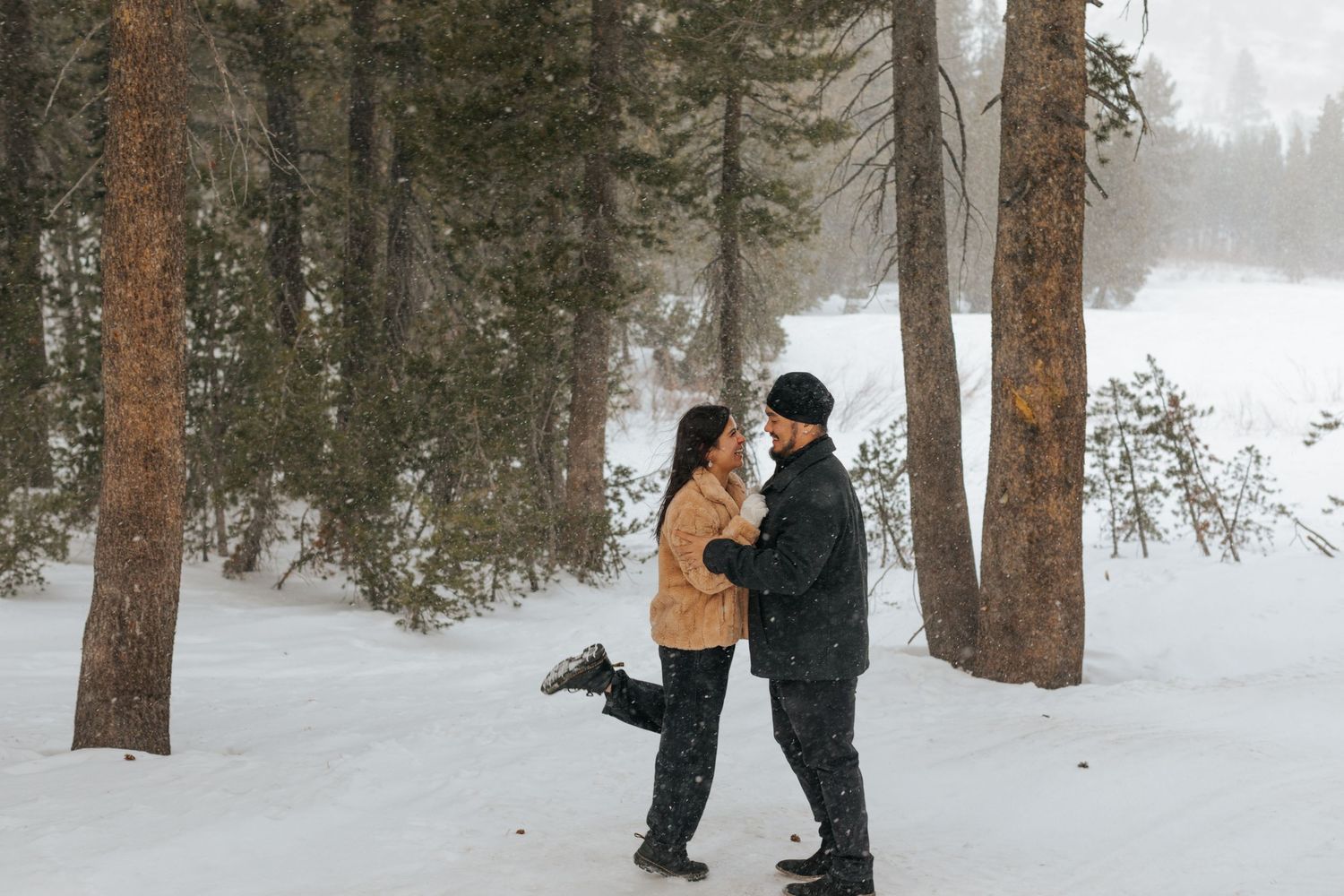 Winter engagement photoshoot in snow-covered forest with tall pine trees.