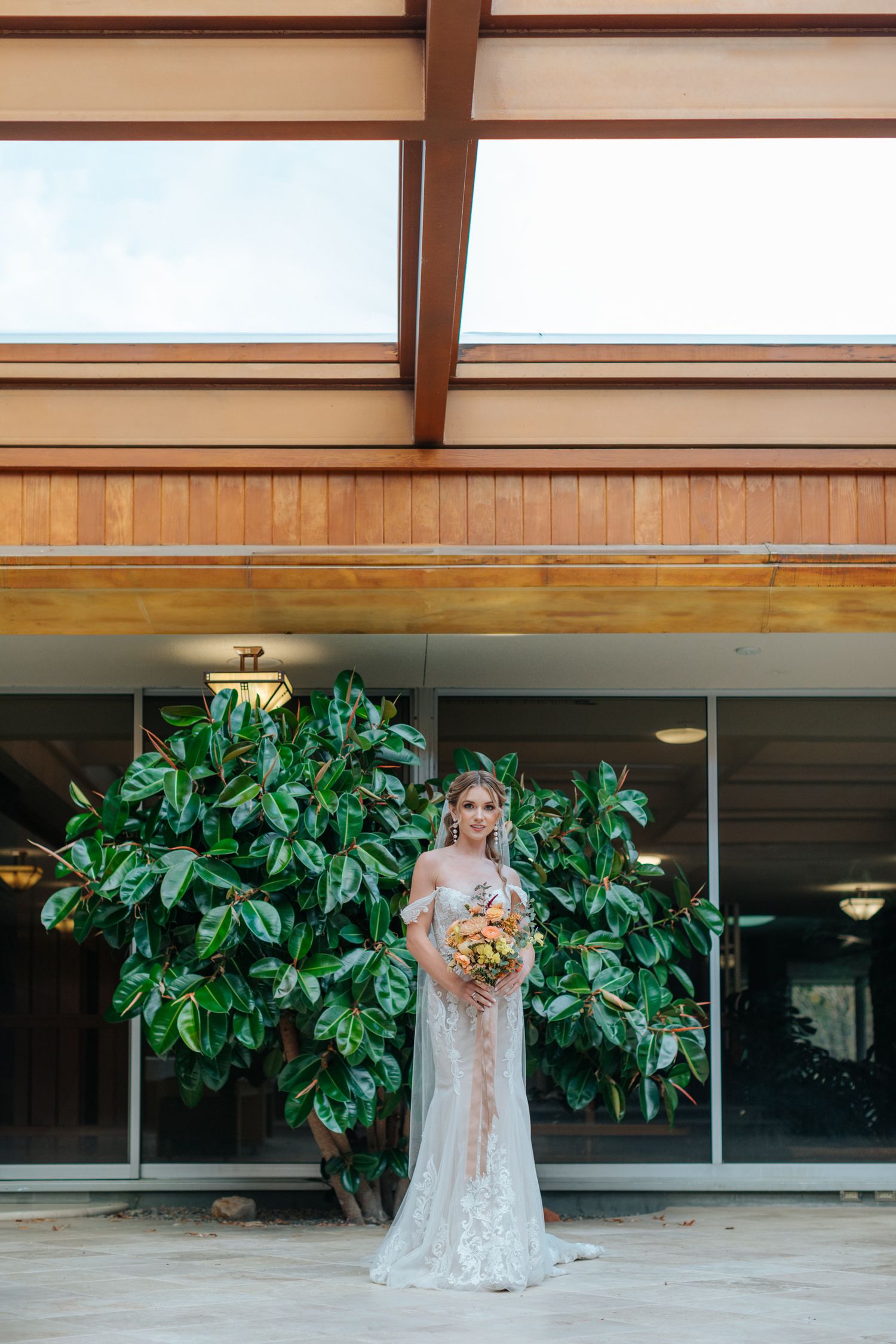 Bride in lace wedding gown holding floral bouquet standing before lush tropical plants at modern venue.