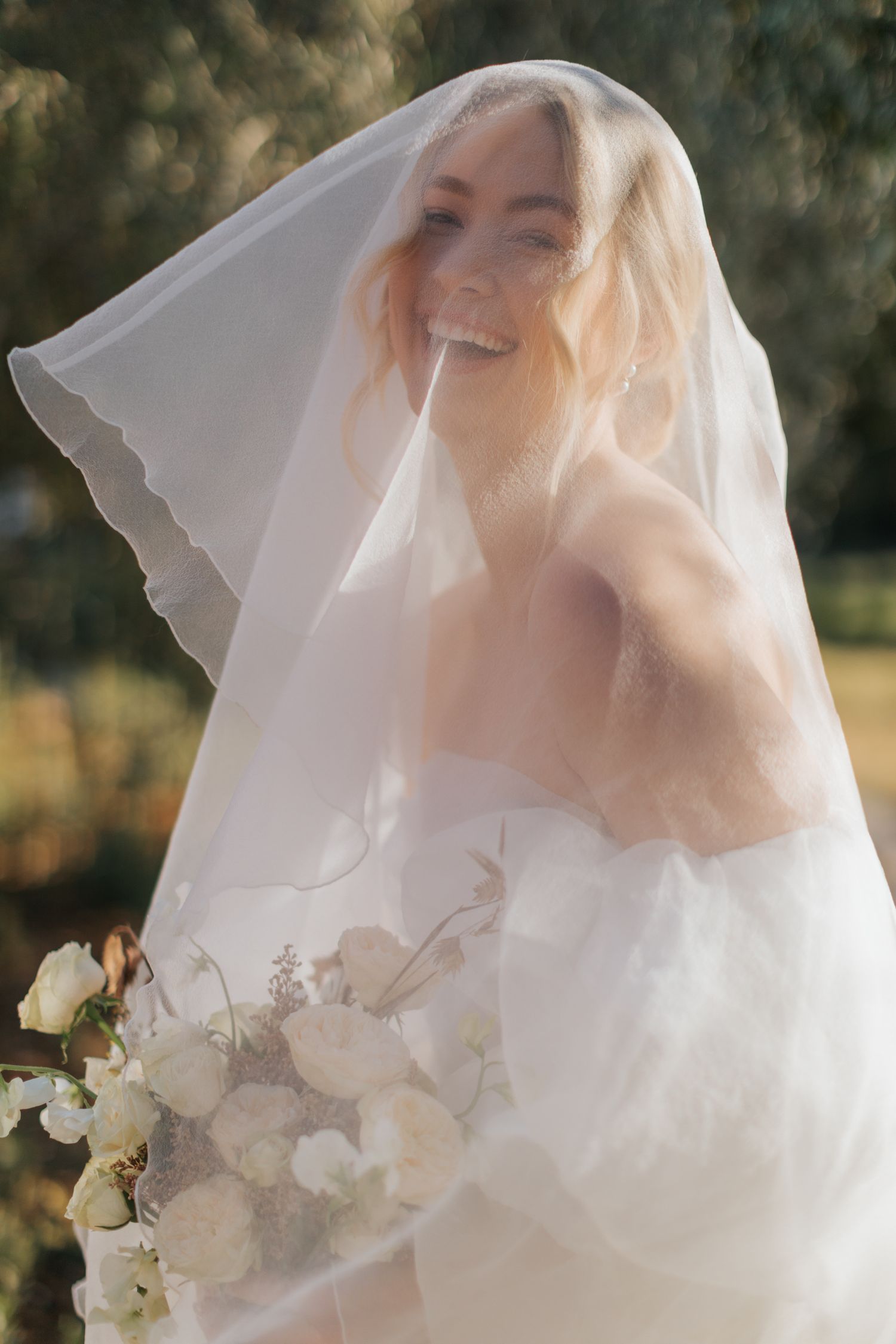 A bride in a white wedding gown and veil holds a delicate bouquet of white flowers in soft sunlit outdoor setting.