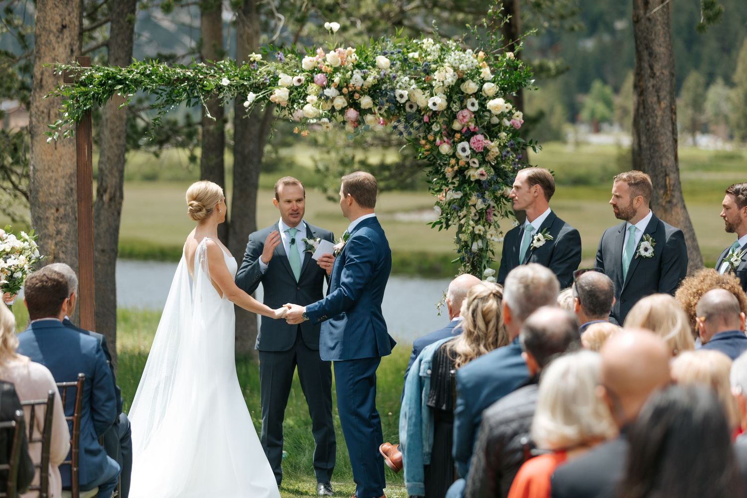 Outdoor wedding ceremony in a pine forest setting with floral arch and seated guests.