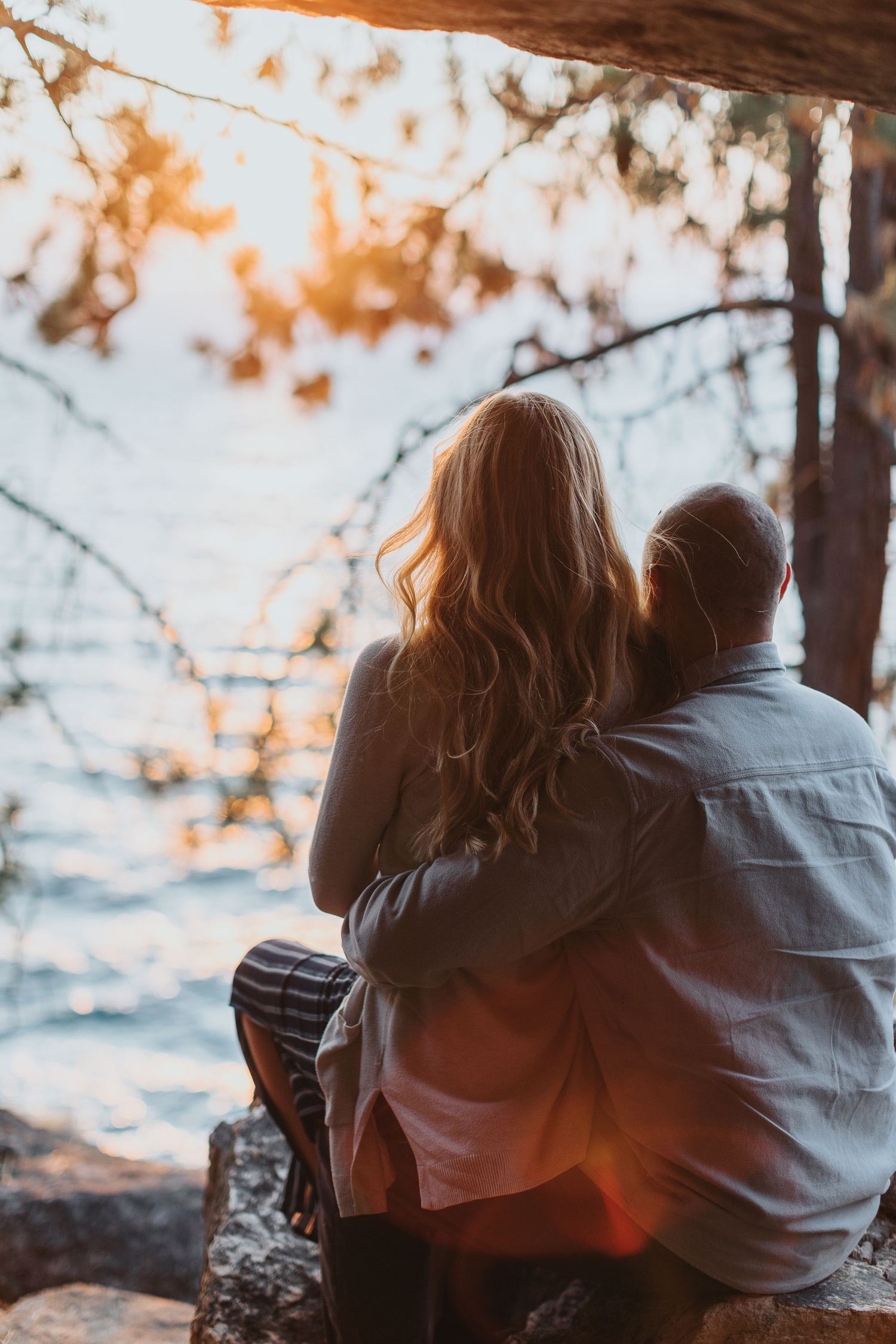 Silhouetted couple watches sunset over ocean through pine trees.