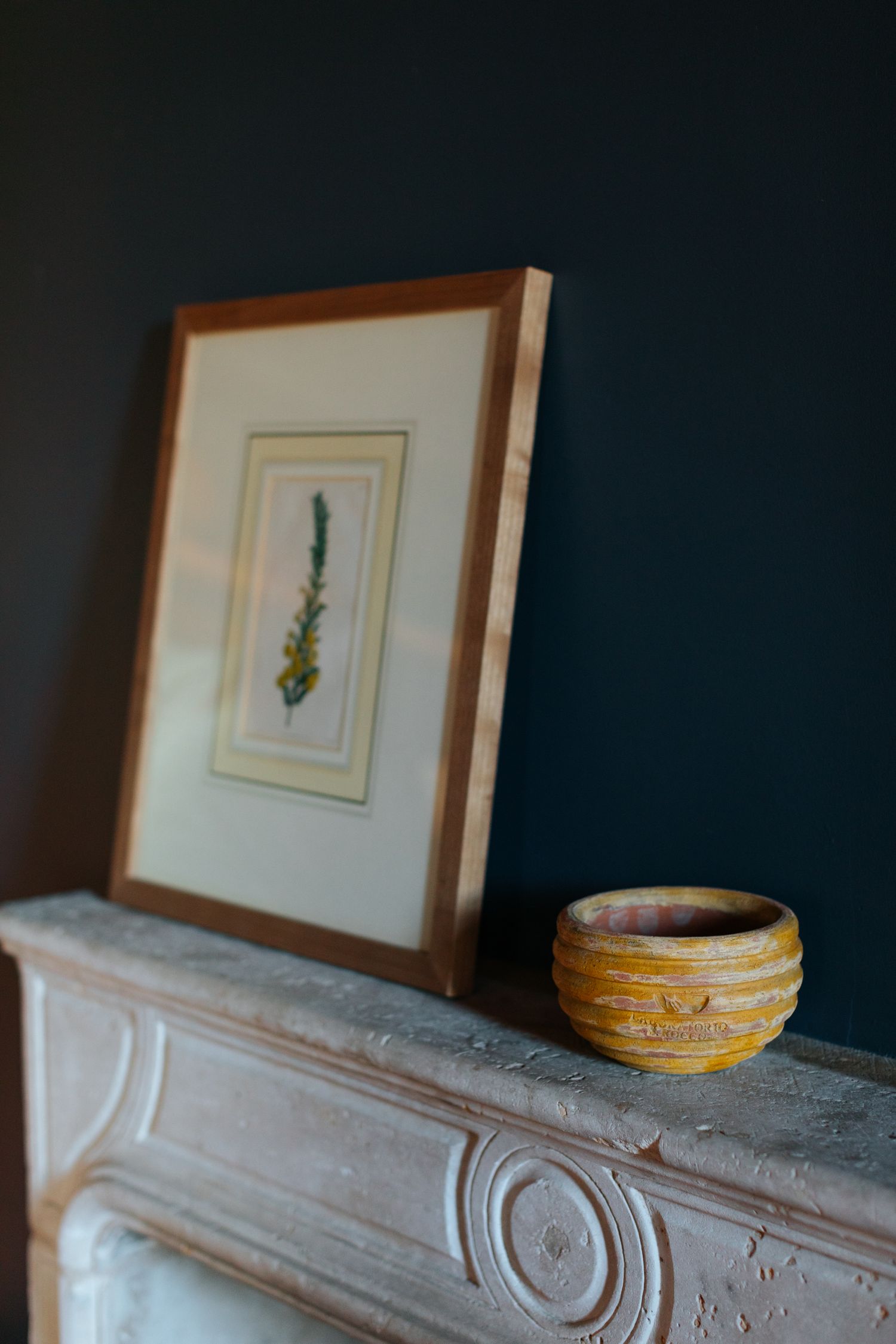 Vintage ornate mantelpiece with framed artwork and decorative bowl against dark wall.