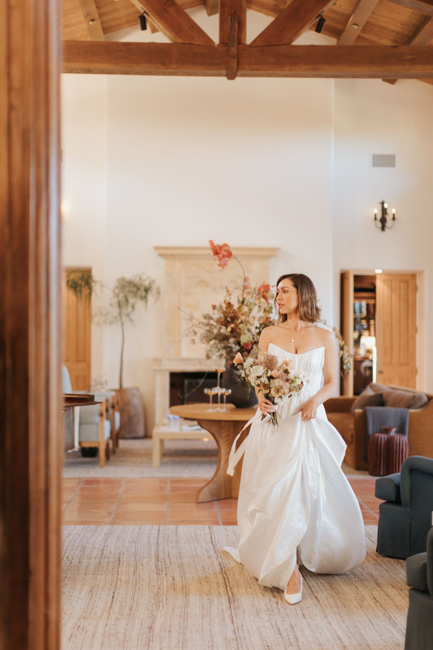 A bride in a flowing white gown holds a romantic bouquet of dried flowers in an elegant room with exposed wooden beams.