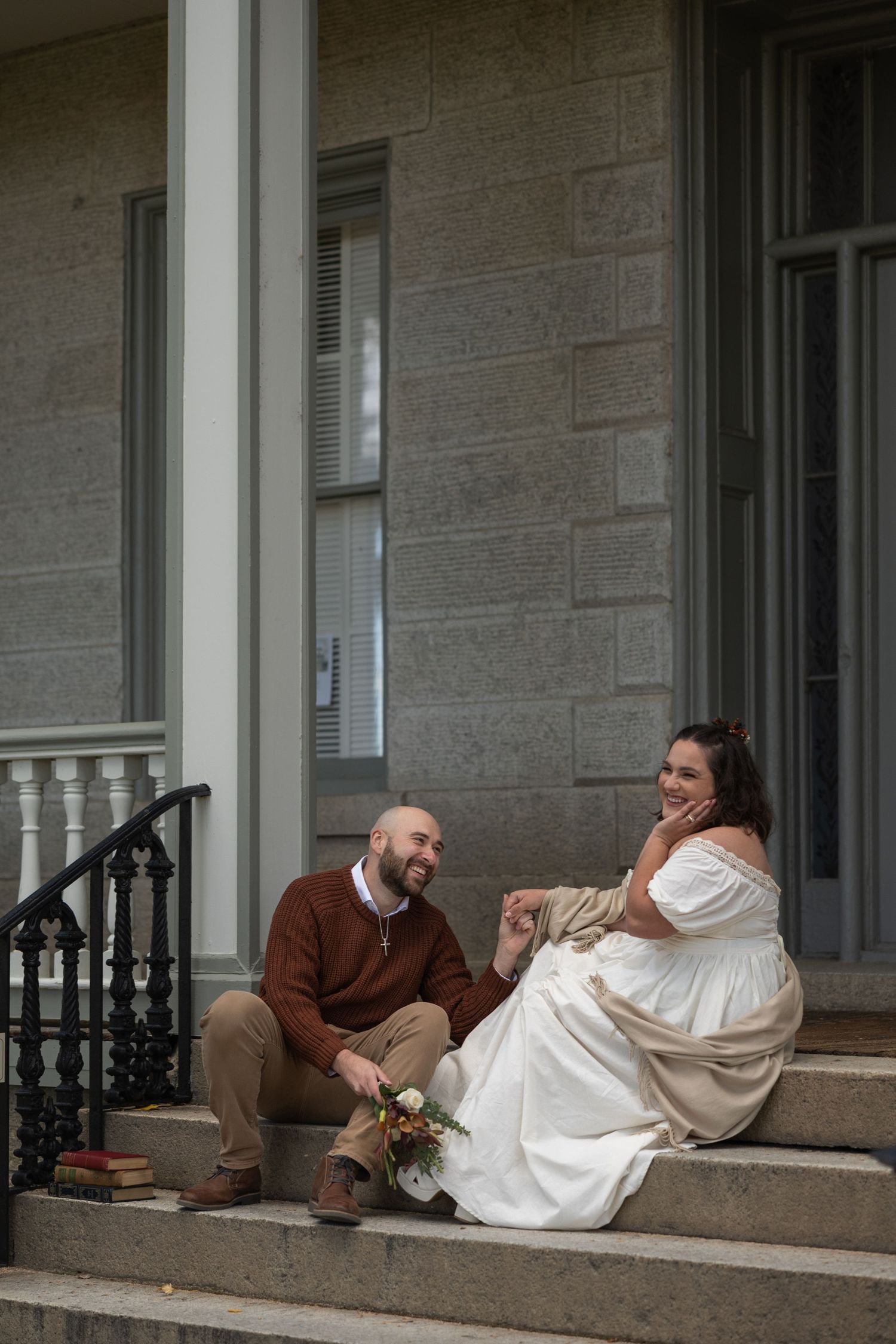 A couple sits together on stone steps of a building entrance, sharing a joyful moment in wedding attire.