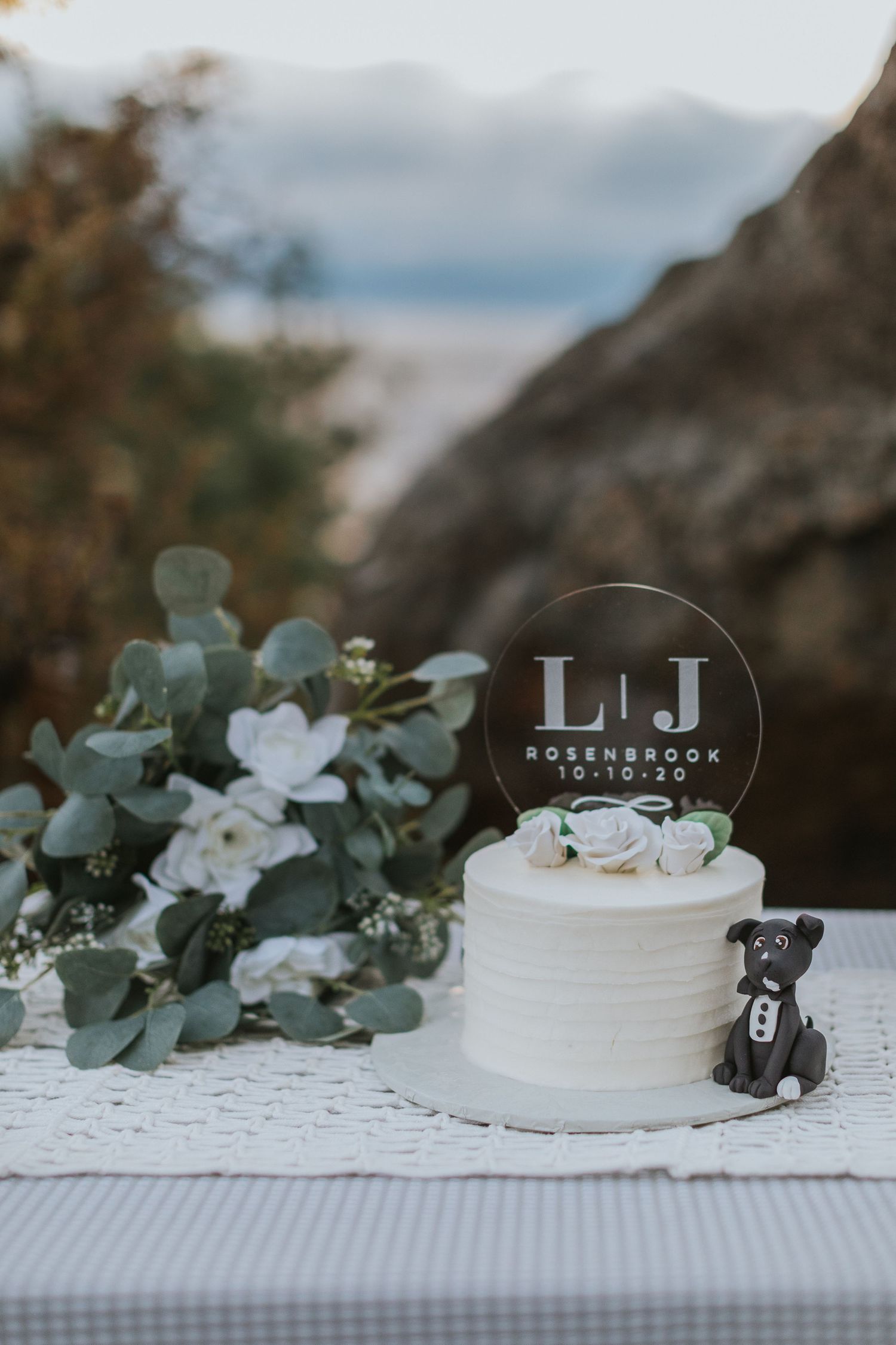 White wedding cake decorated with eucalyptus and flowers on a textured tablecloth.