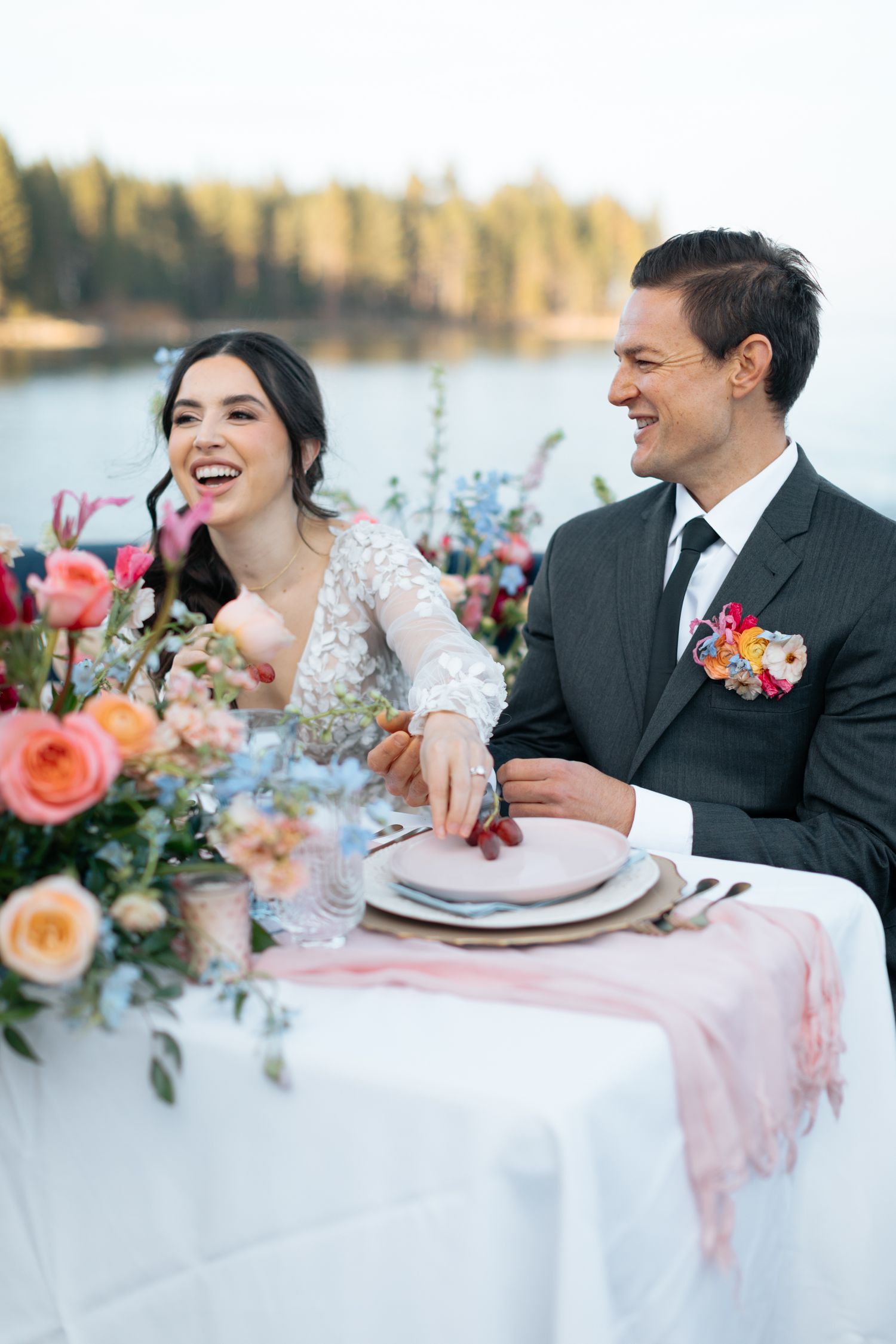 Laughing bride and groom seated at floral-decorated lakeside reception table, groom wearing colorful boutonniere.