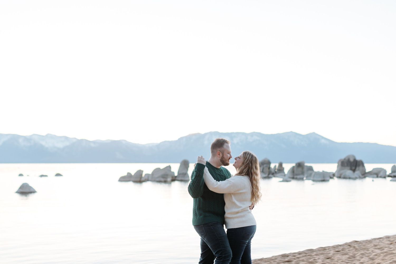 Beautiful lakeside beach scene with mountains and rocks visible in the distance.
